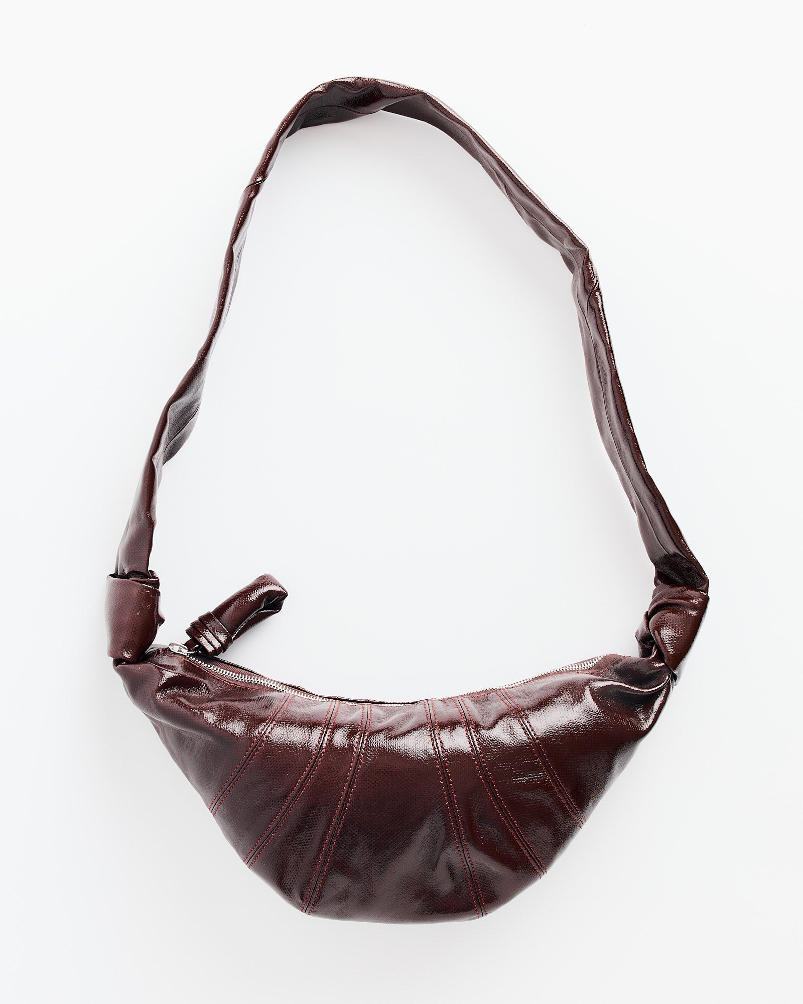 The Lemaire Small Croissant Bag in Chocolate Fondant is a dark brown, shiny bag with a wide curved strap and zipper closure, made of coated cotton canvas and shown on a white background.