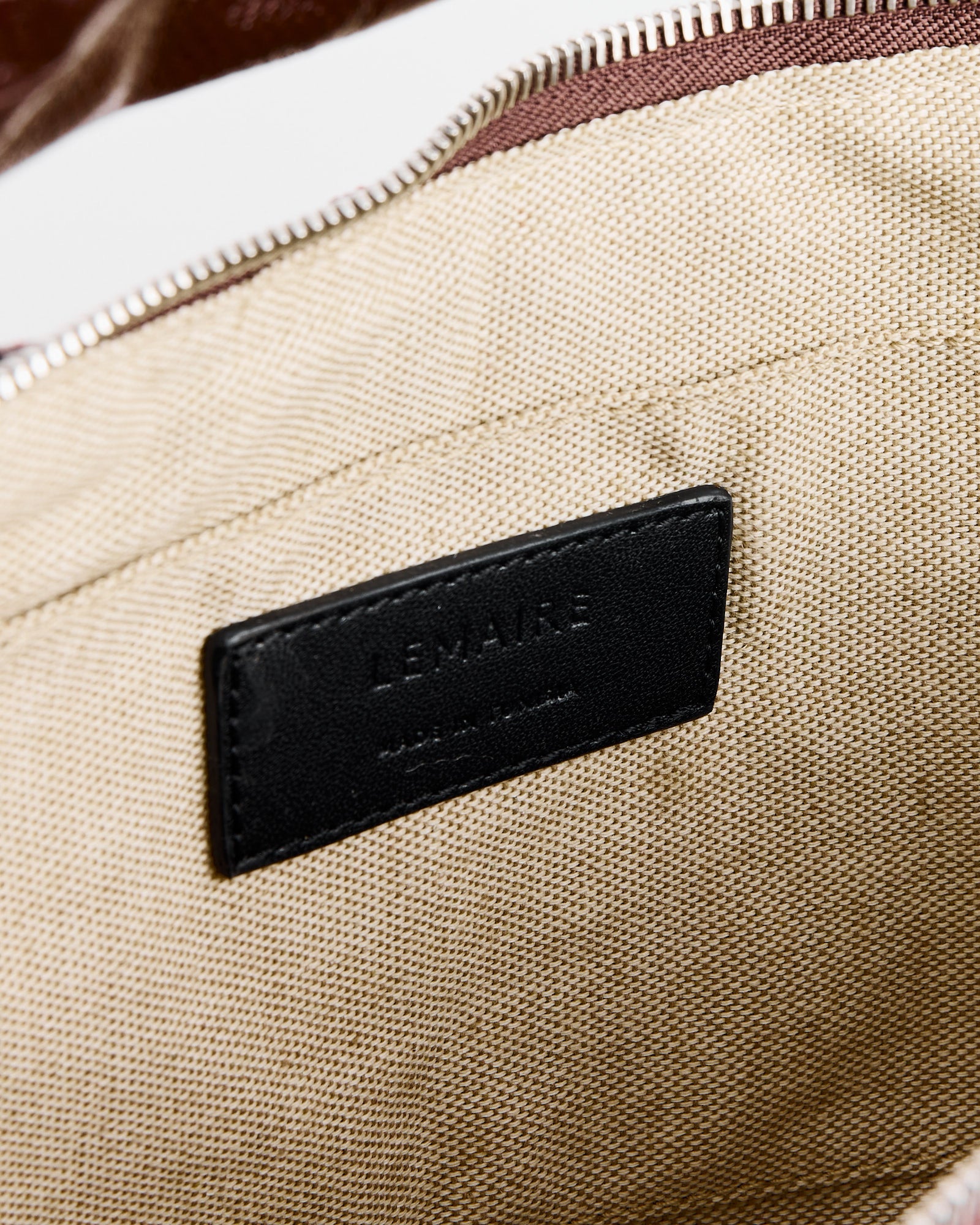 Close-up of the beige coated cotton canvas interior of the Lemaire Small Croissant Bag in Chocolate Fondant, featuring a black leather "LEMAIRE MADE IN SPAIN" label, with a silver zipper and brown trim visible at the top.