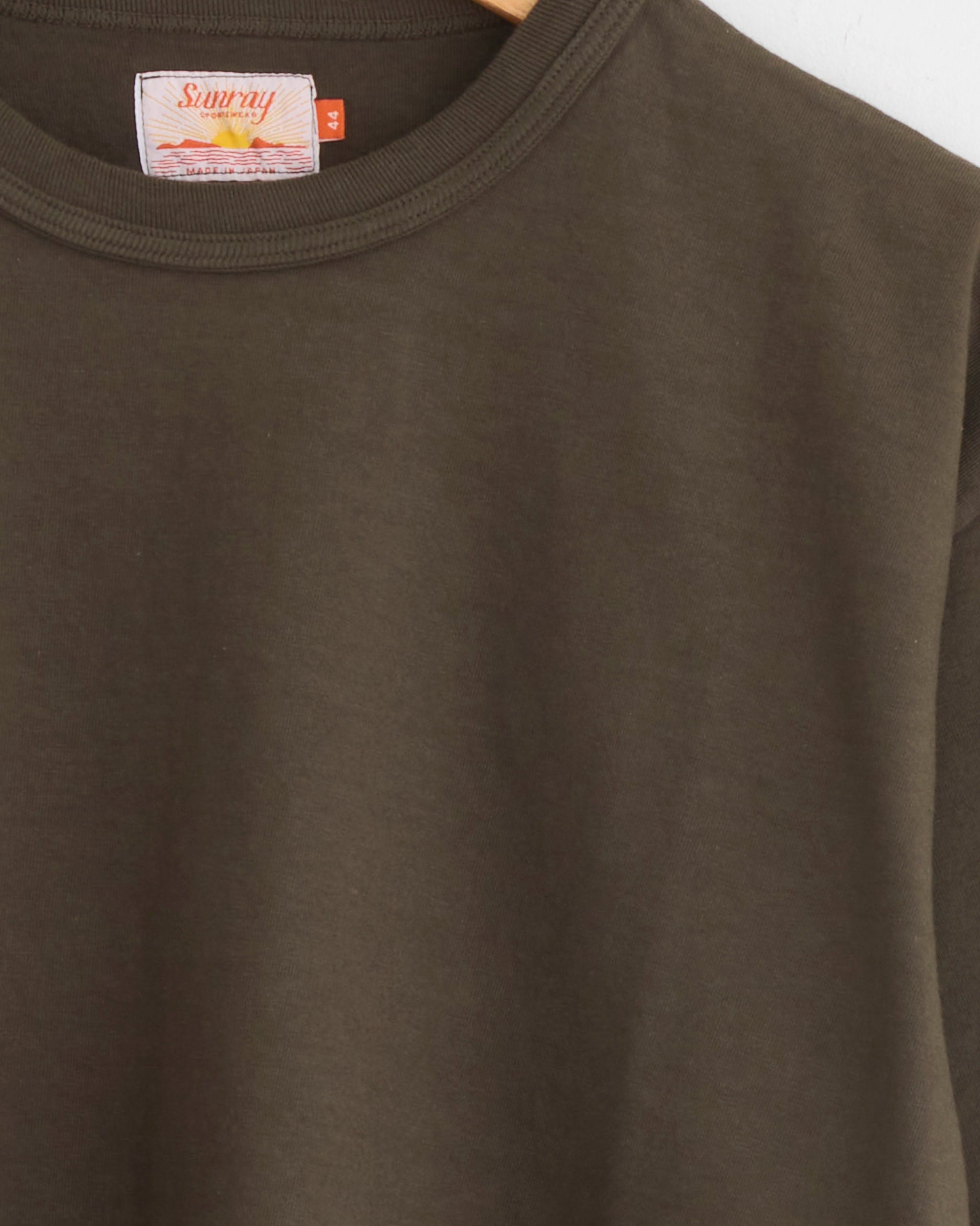 A close-up of the Makaha Japanese Jersey T-Shirt in Black Olive by Sunray hangs on a wooden hanger, highlighting the upper section and featuring a visible Sunray brand tag at the neckline.