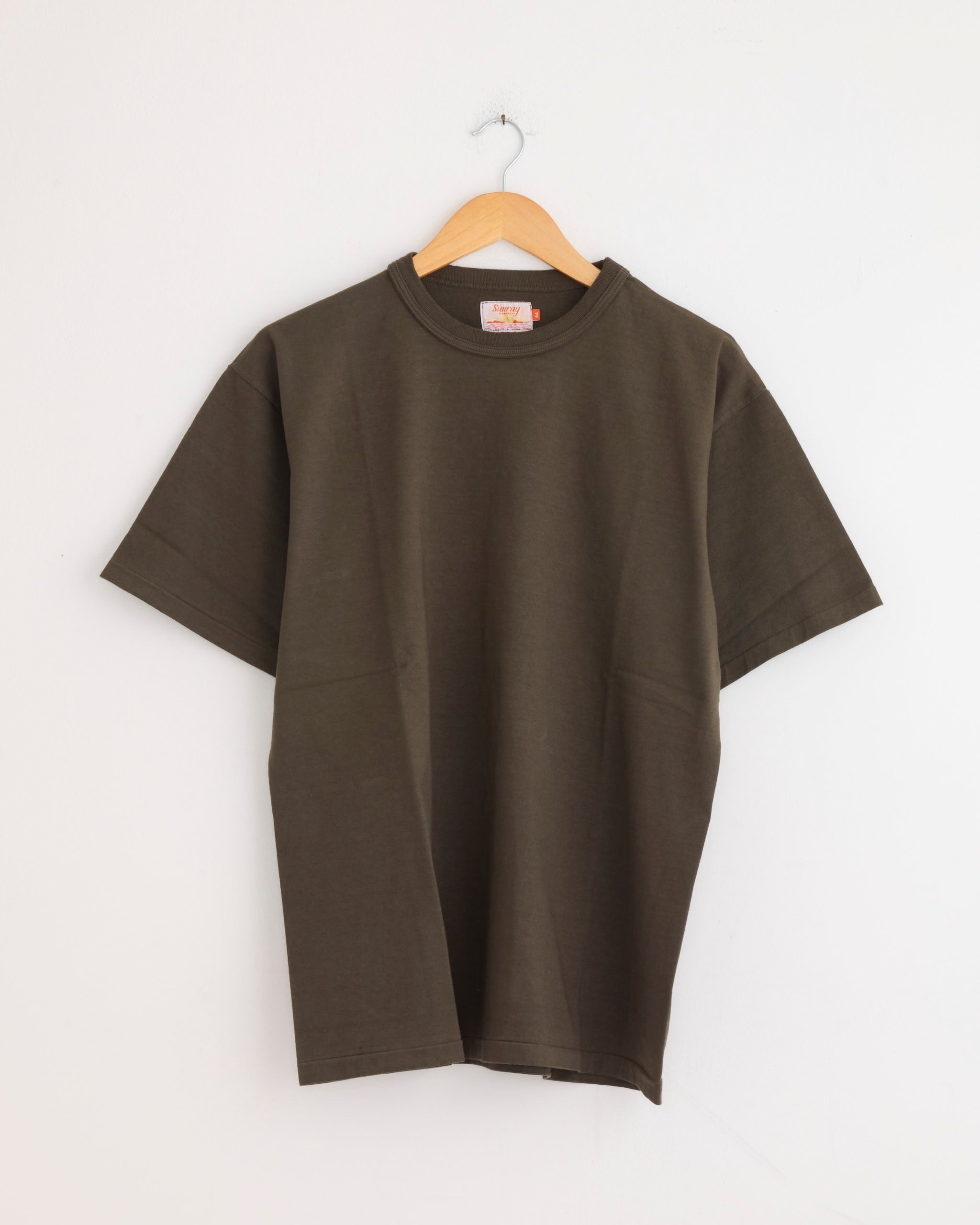 The Sunray Makaha Japanese Jersey T-Shirt in Black Olive is displayed on a wooden hanger against a white wall.