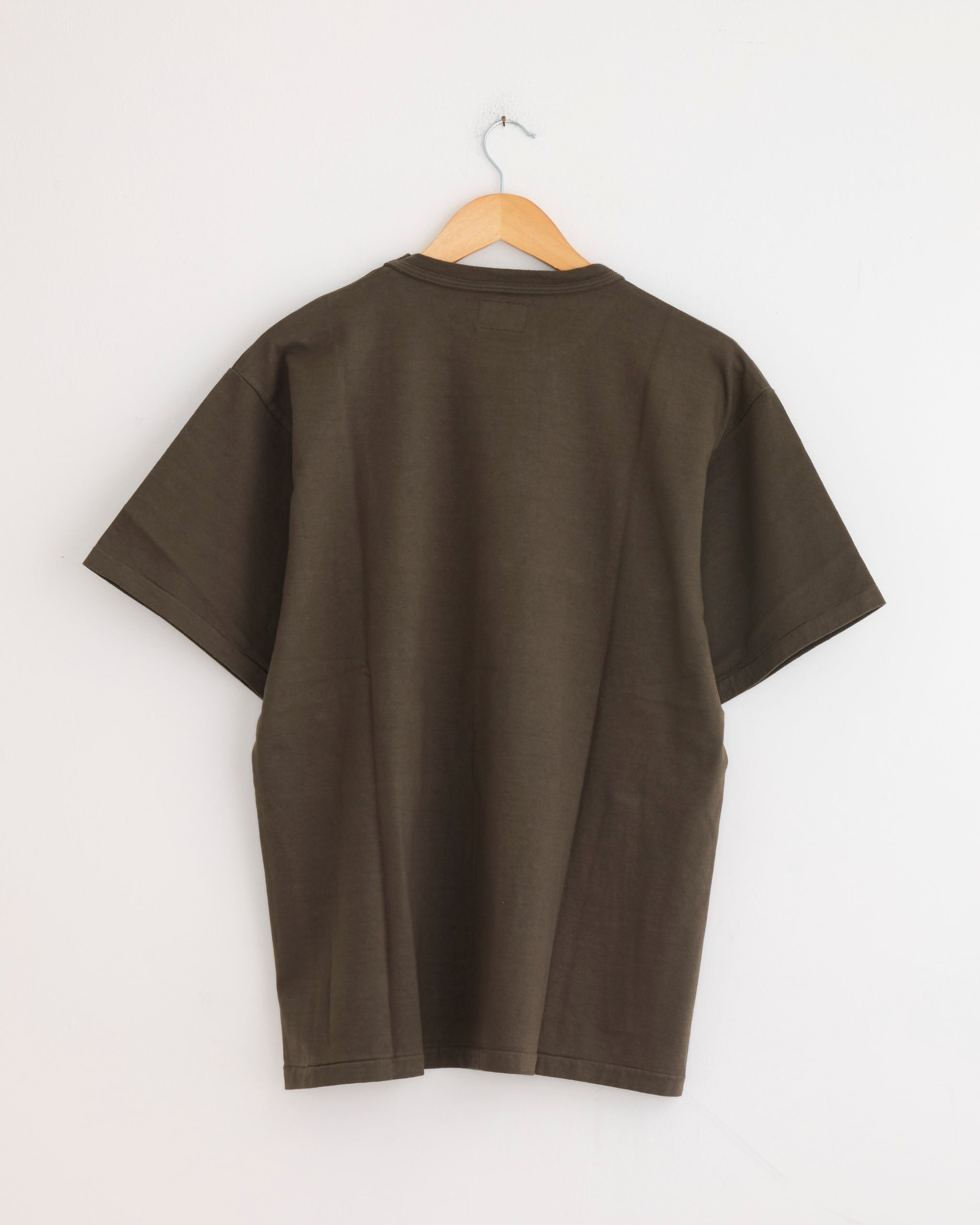 The Sunray Makaha Japanese Jersey T-Shirt in Black Olive, made from heavyweight fabric, is displayed from the back on a wooden hanger against a white wall.