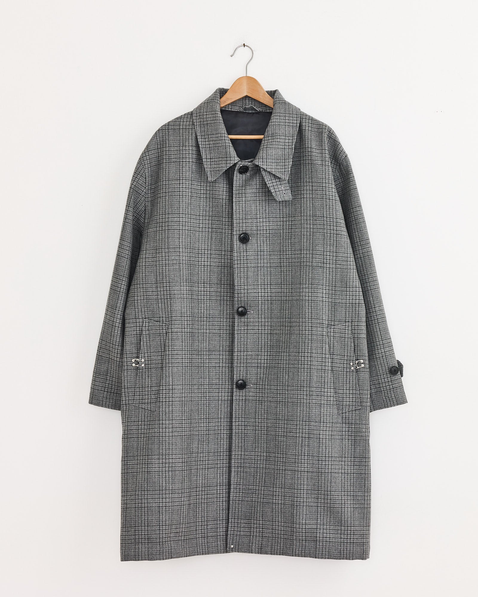 The mfpen Client Coat in Grey Glen Check, made from Italian deadstock wool, features a collar, button-down front, and two side pockets. It hangs on a wooden hanger against a white wall.