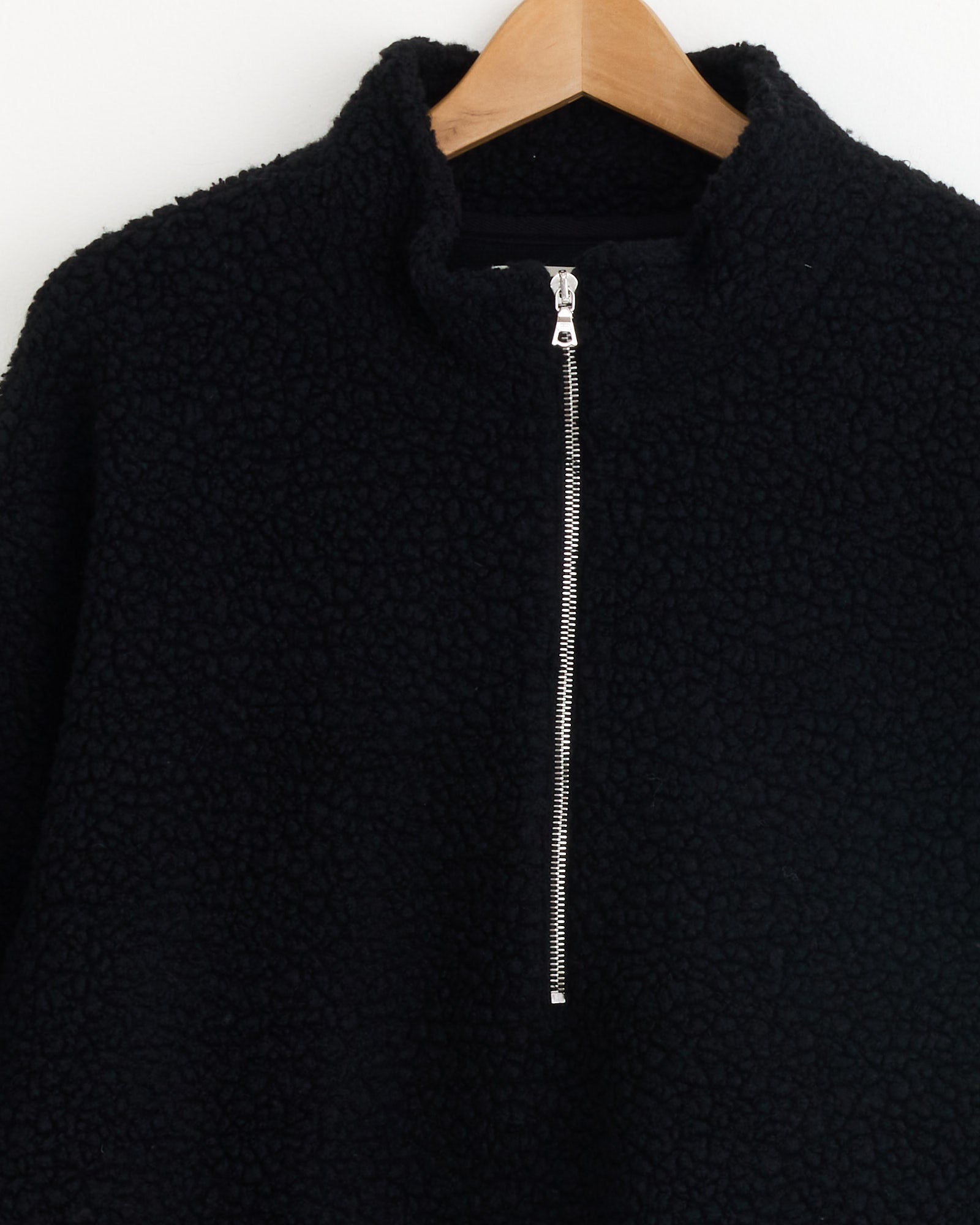 The mfpen Common Half Zip Sweater in Black, with textured sherpa-like fleece, a silver half-zipper, and stand-up collar, is shown close-up on a wooden hanger against a white background.