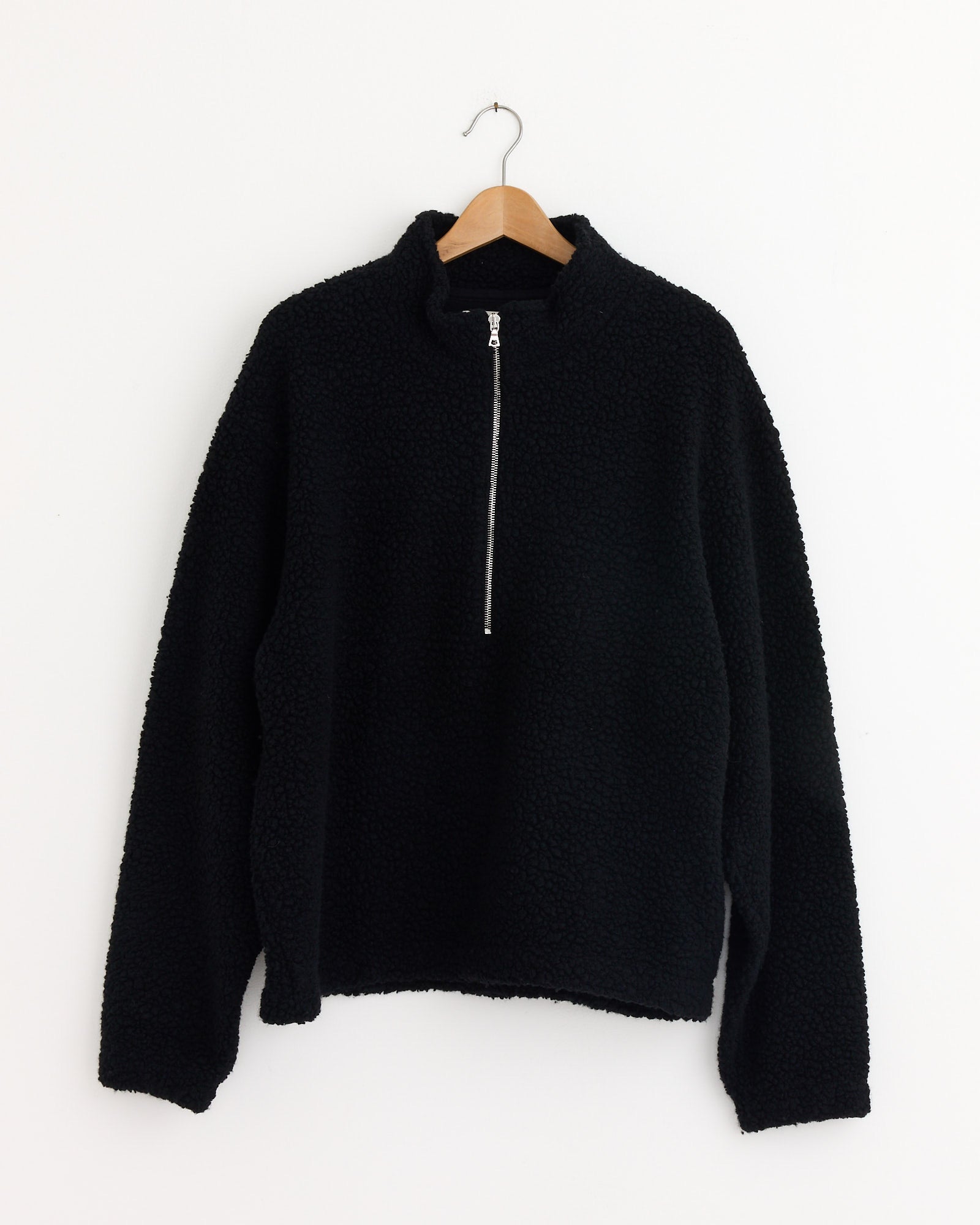 The mfpen Common Half Zip Sweater in Black, featuring a quarter-zip front, long sleeves, and a high collar, hangs on a wooden hanger against a white wall.