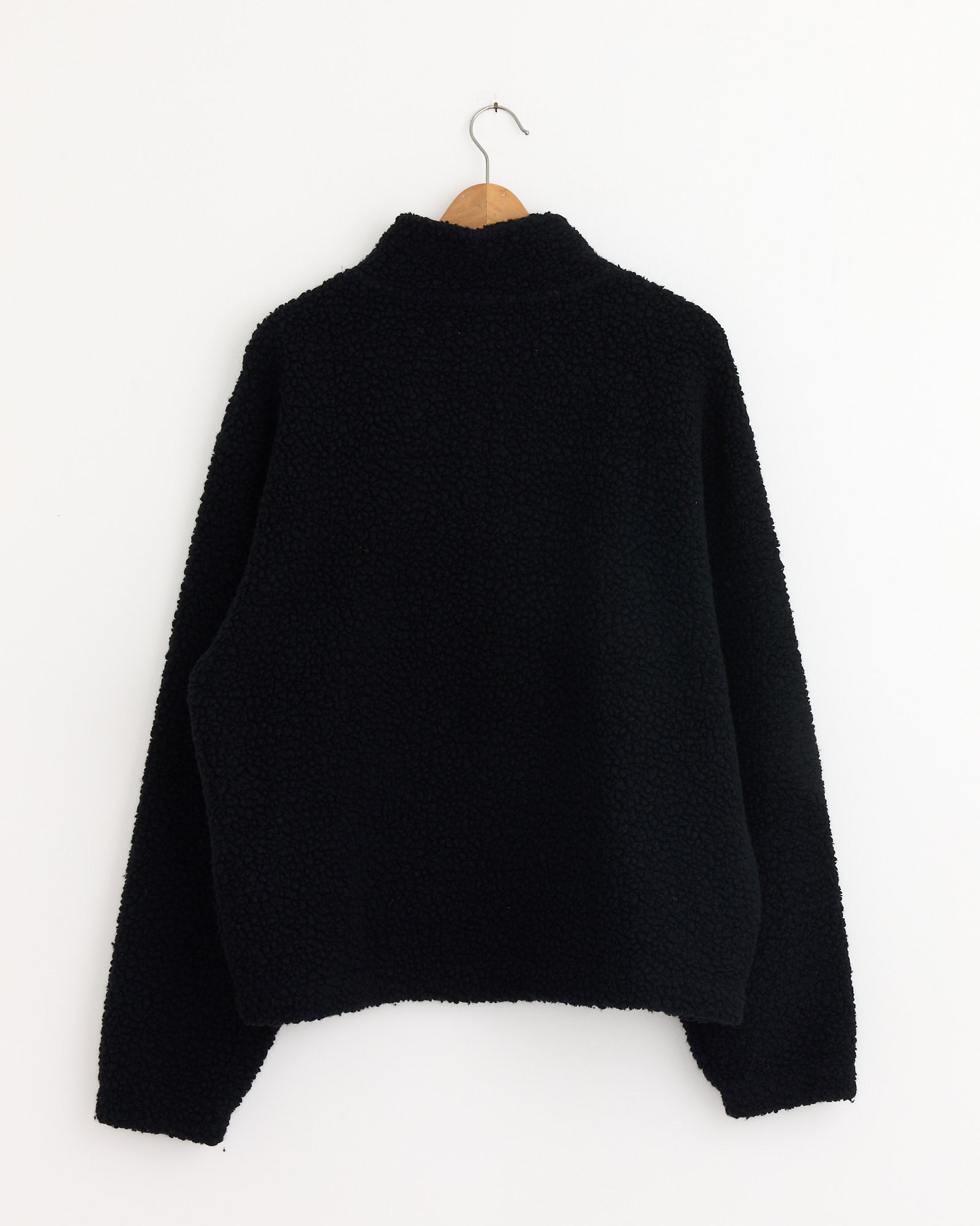 The mfpen Common Half Zip Sweater in Black, with a textured finish, is displayed from the back on a wooden hanger against a plain white wall.