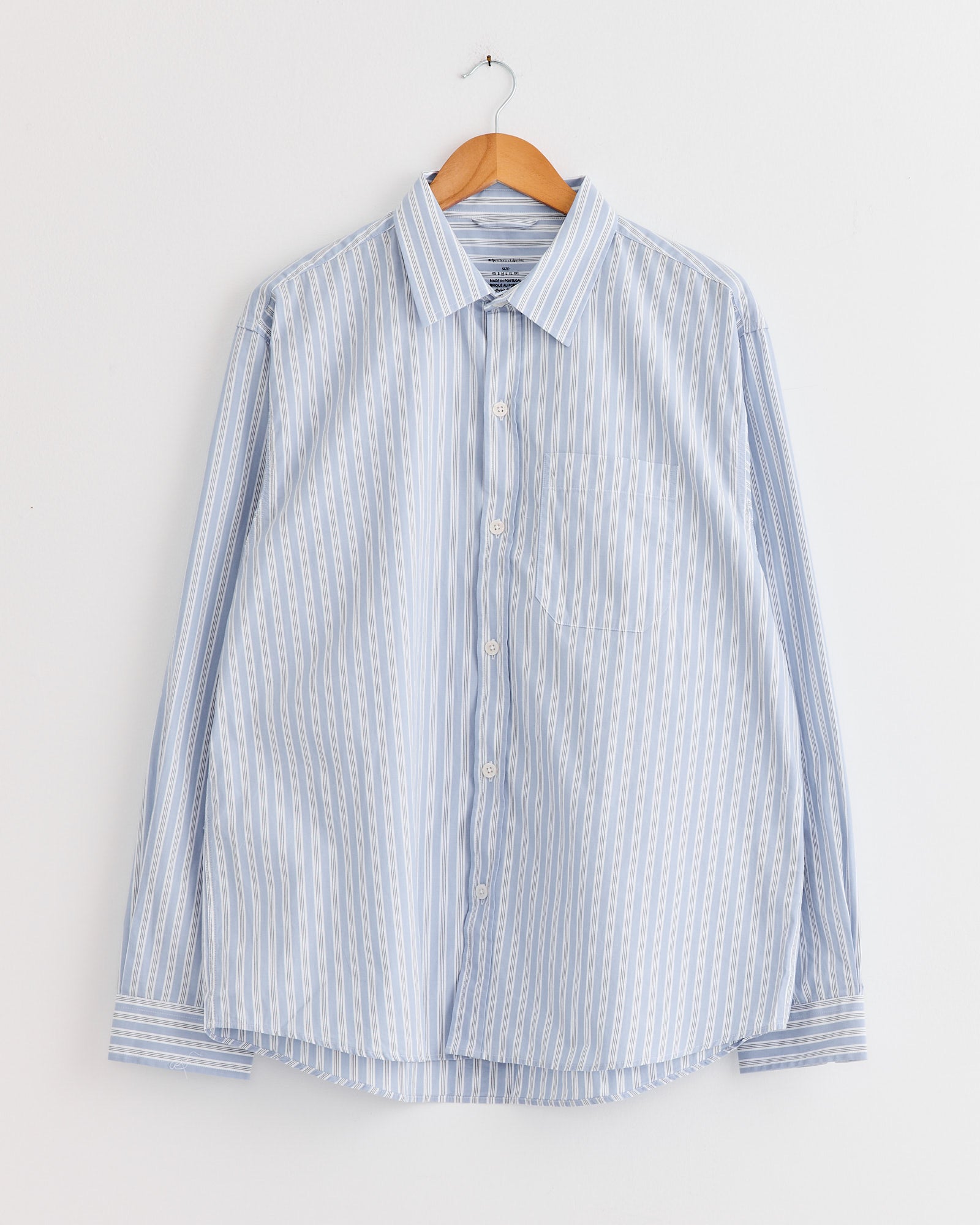 The mfpen Distant Shirt in Finance Stripe, a light blue and white vertically striped long-sleeve button-up made from organic cotton, hangs on a wooden hanger against a plain white wall.