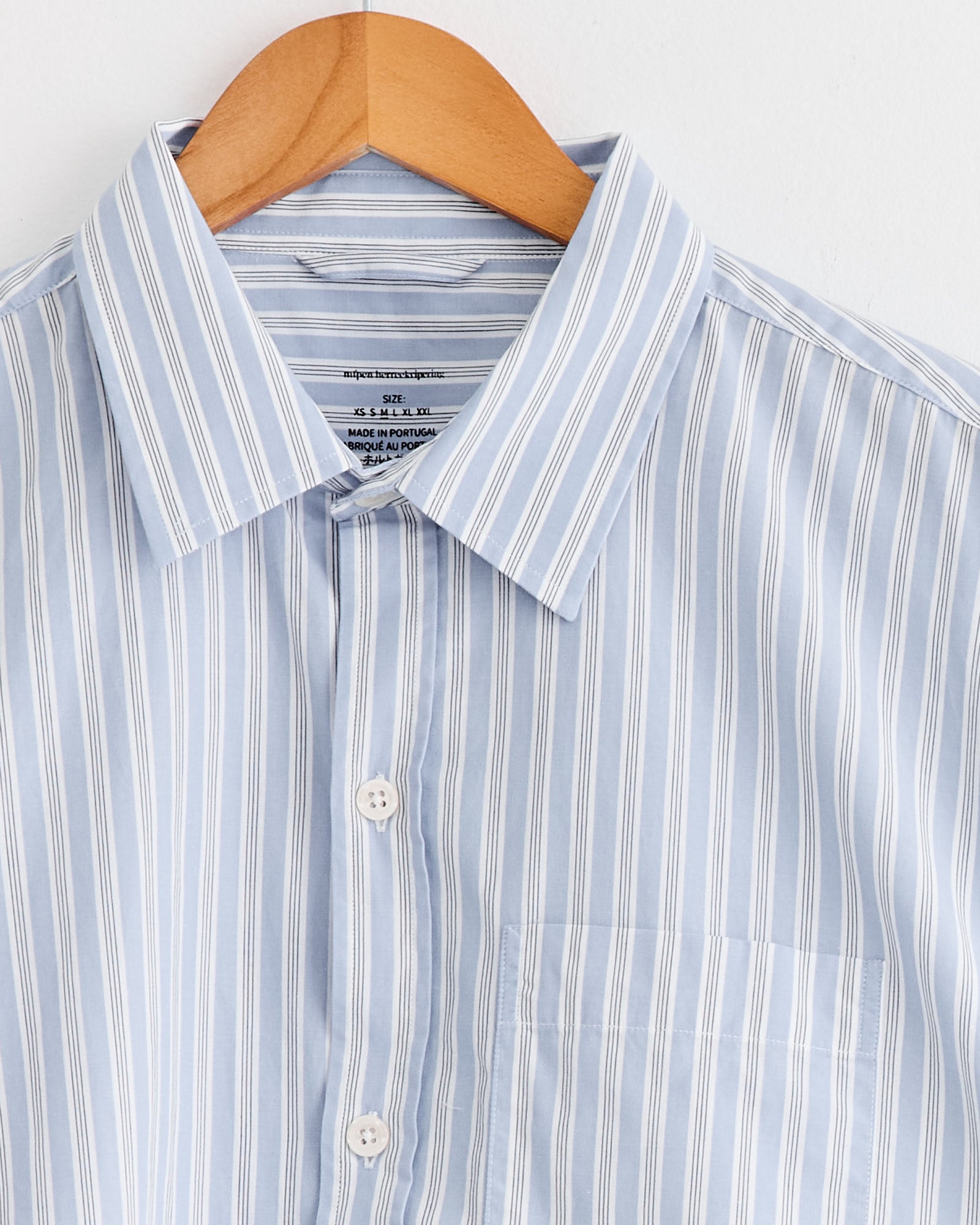 The mfpen Distant Shirt in Finance Stripe is a light blue collared organic cotton shirt with white vertical stripes, buttoned up and displayed on a wooden hanger against a white background.