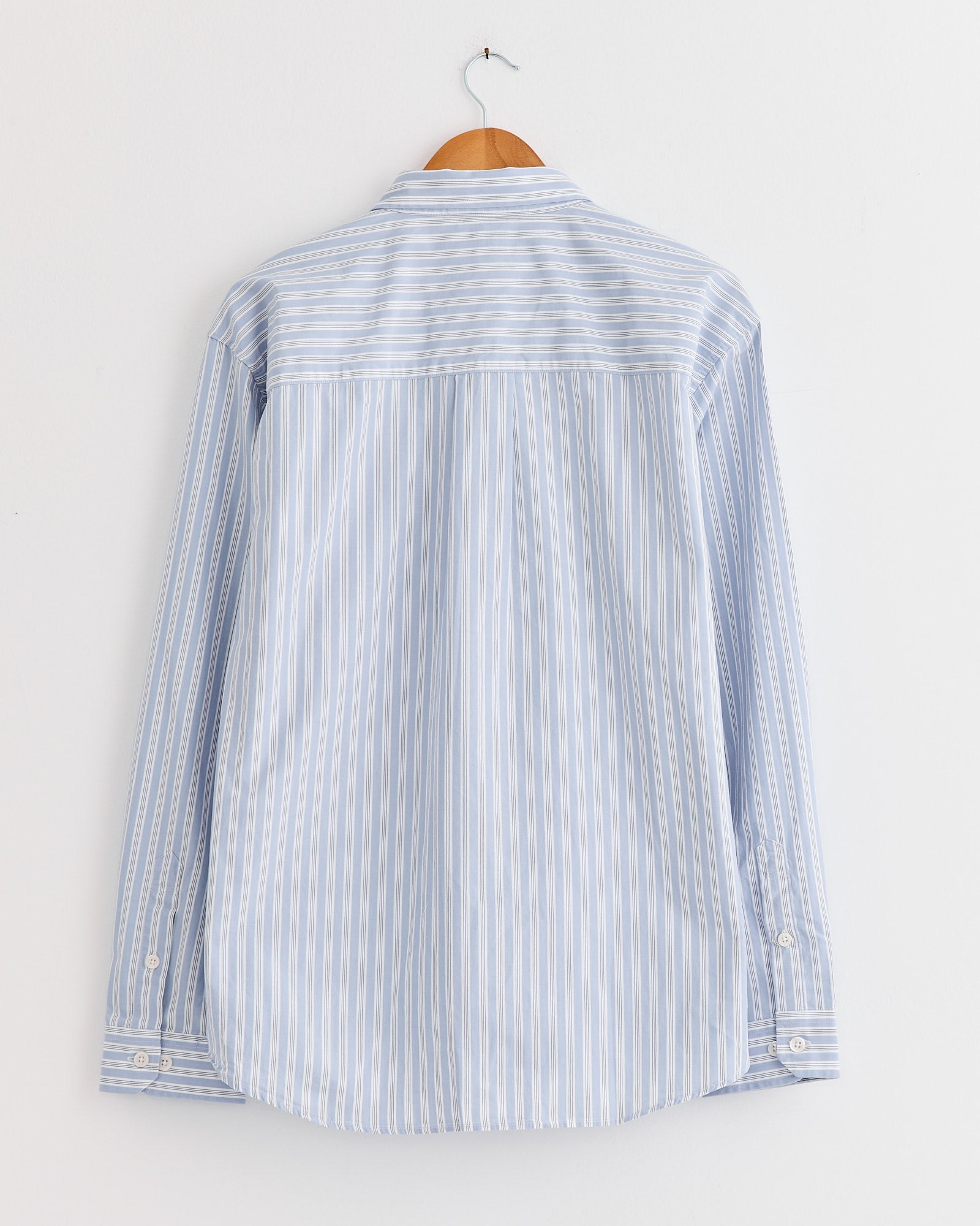 The mfpen Distant Shirt in Finance Stripe, featuring light blue and white vertical stripes, hangs on a wooden hanger against a white wall, shown from the back.