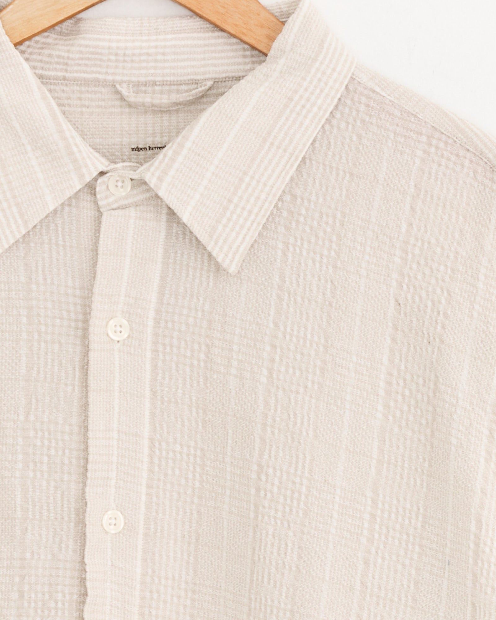 A close-up of the mfpen Exact Shirt in Dusty Seersucker Check on a wooden hanger, highlighting the collar, buttons, and part of the sleeve against a white background.
