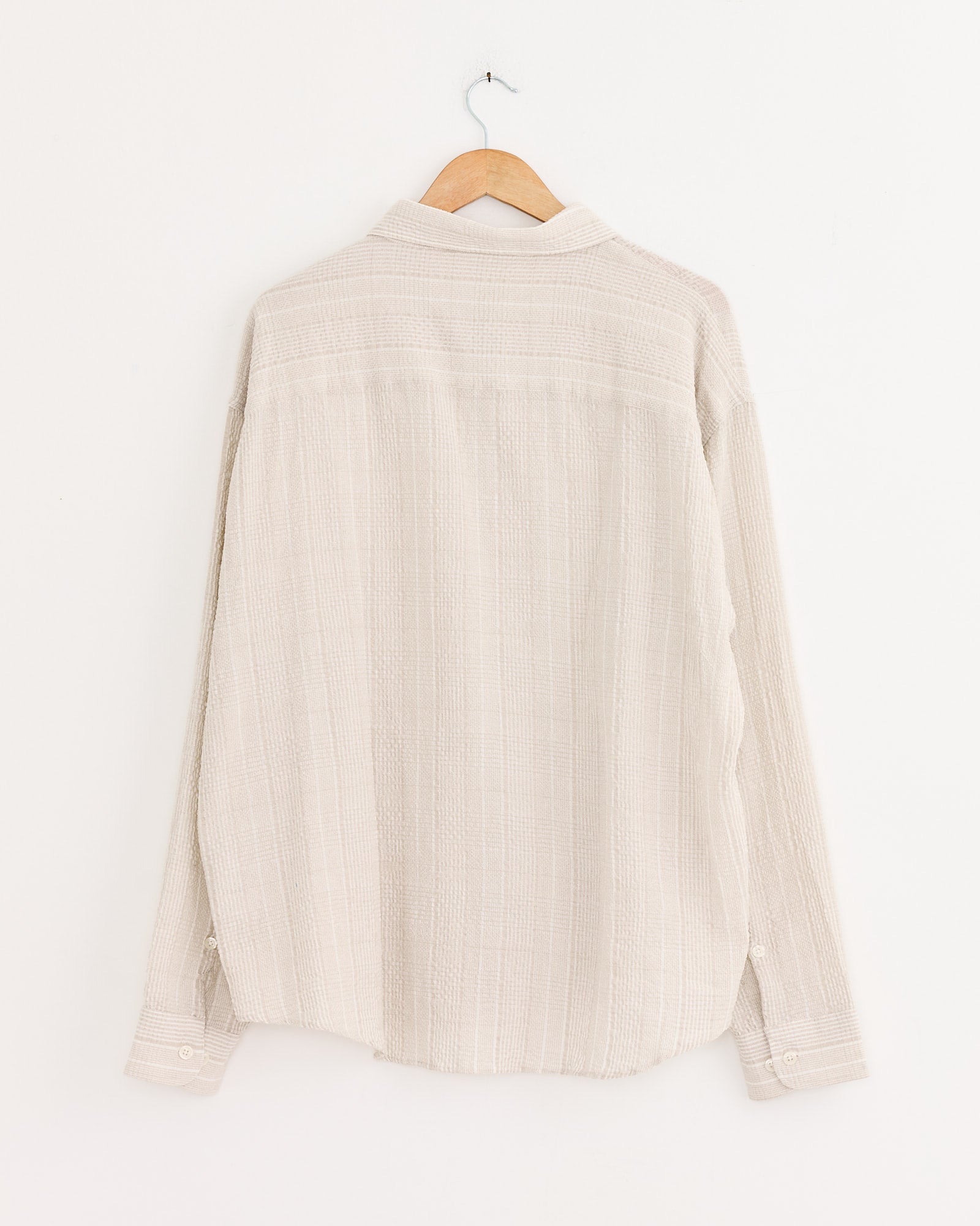 The mfpen Exact Shirt in Dusty Seersucker Check, made from recycled cotton, hangs on a wooden hanger against a plain white wall, shown from the back.