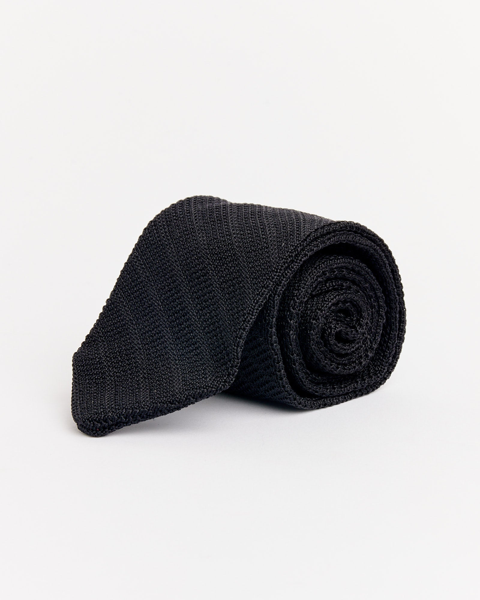 The mfpen Formal Tie in Black Knitted Stripe, made in Italy, is shown rolled up against a plain white background.