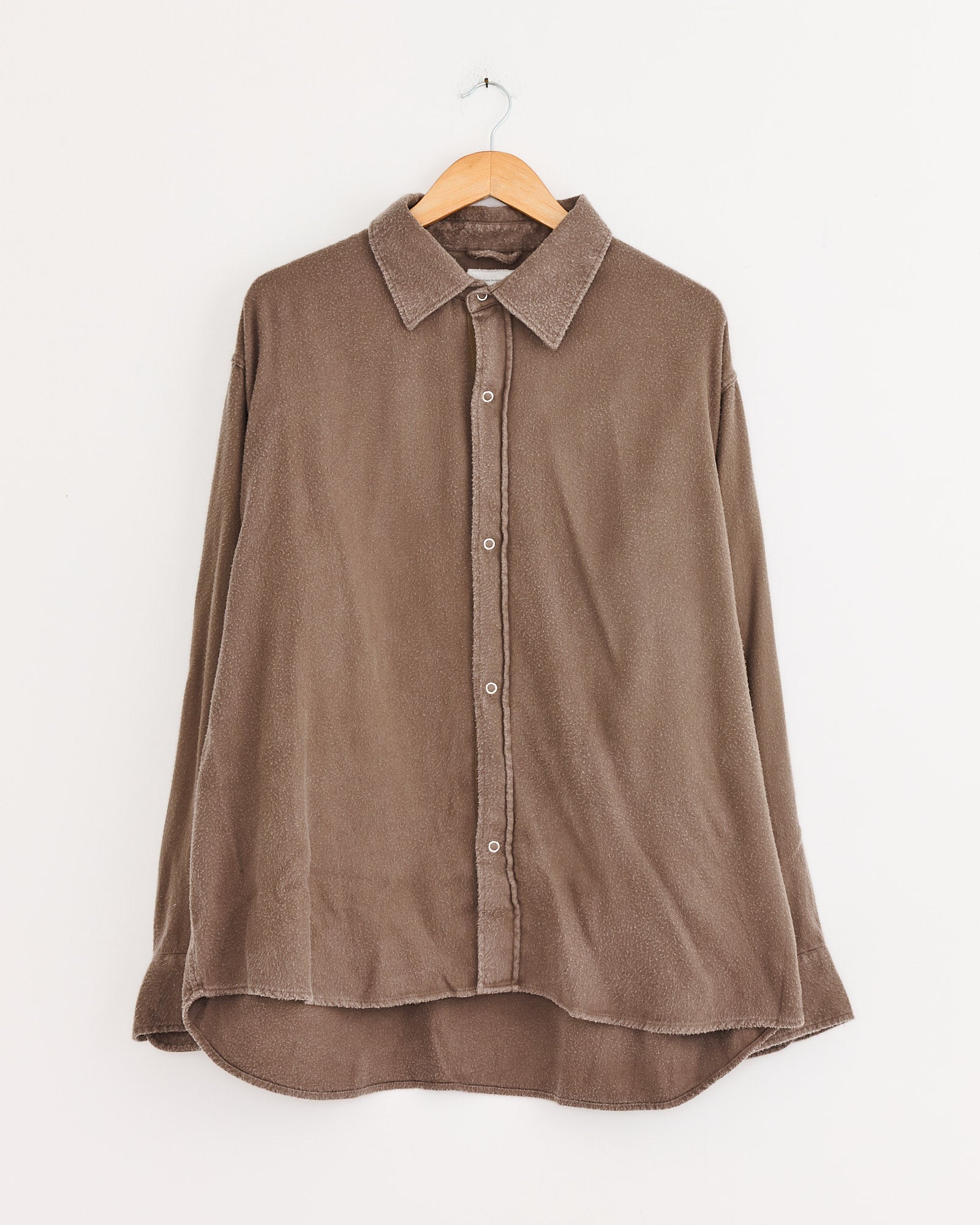 The mfpen Gambler Shirt in Dark Taupe Fuzz, an oversized corduroy style with long sleeves, collar, buttons, and a curved hem, is displayed on a wooden hanger against a plain white wall.