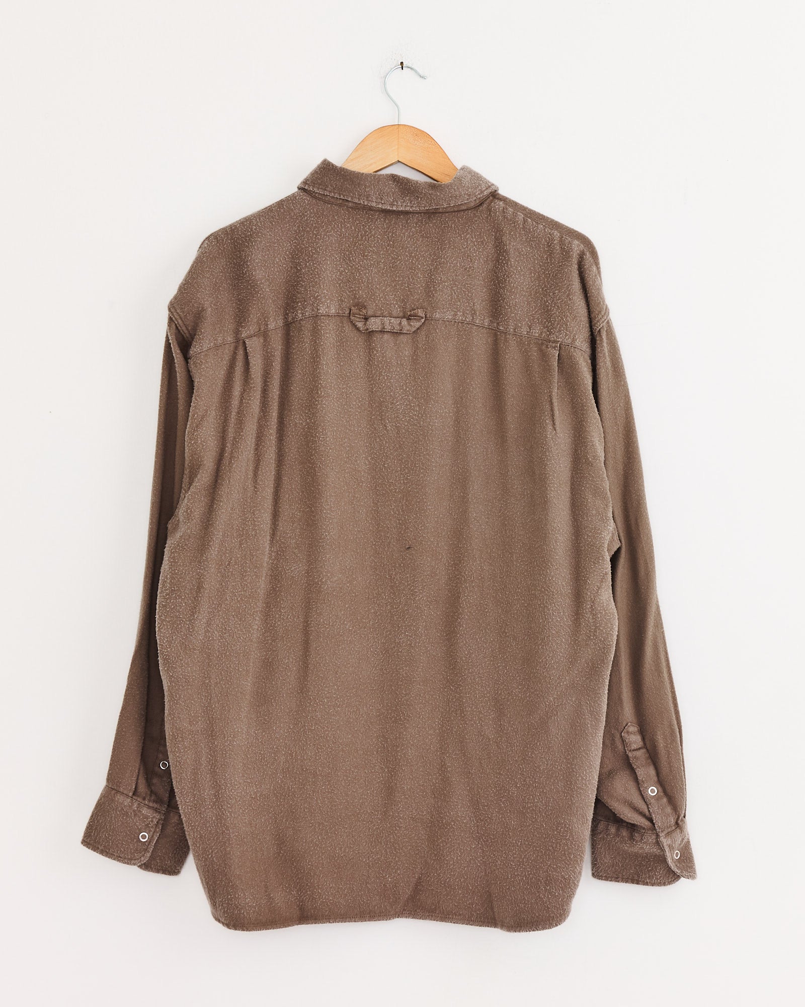 An oversized mfpen Gambler Shirt in Dark Taupe Fuzz hangs on a wooden hanger against a white wall, displayed from the back with a single loop detail below the collar and buttoned cuffs.
