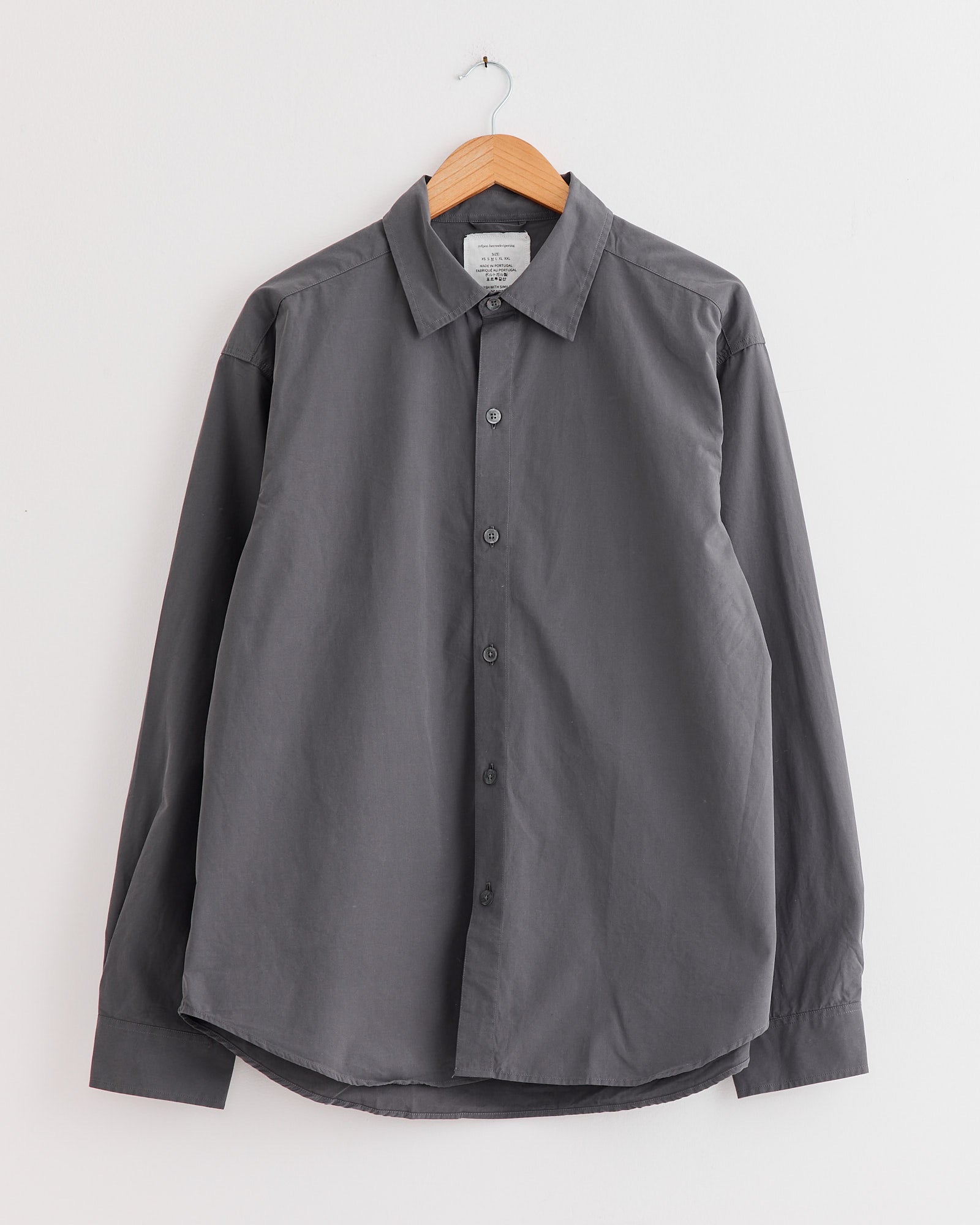 The mfpen Generous Shirt in Mushroom, made of organic cotton with long sleeves and a relaxed fit, hangs on a wooden hanger against a white wall.
