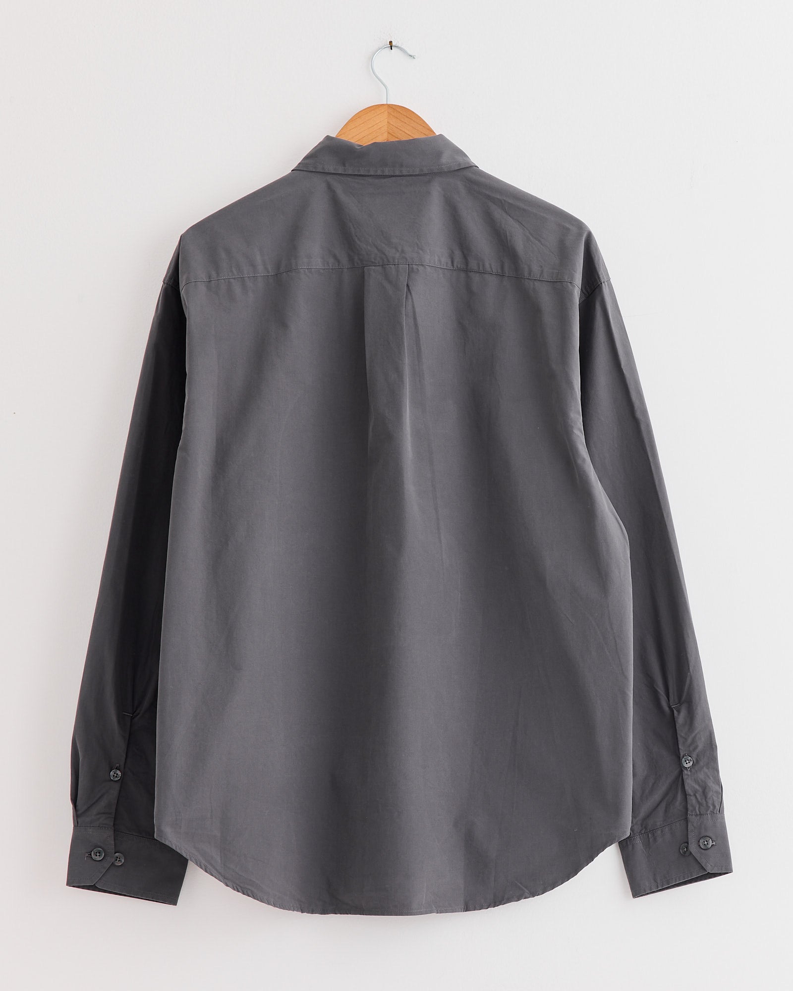 A long-sleeved mfpen Generous Shirt in Mushroom organic cotton with a relaxed fit hangs on a wooden hanger against a white wall, shown from the back.