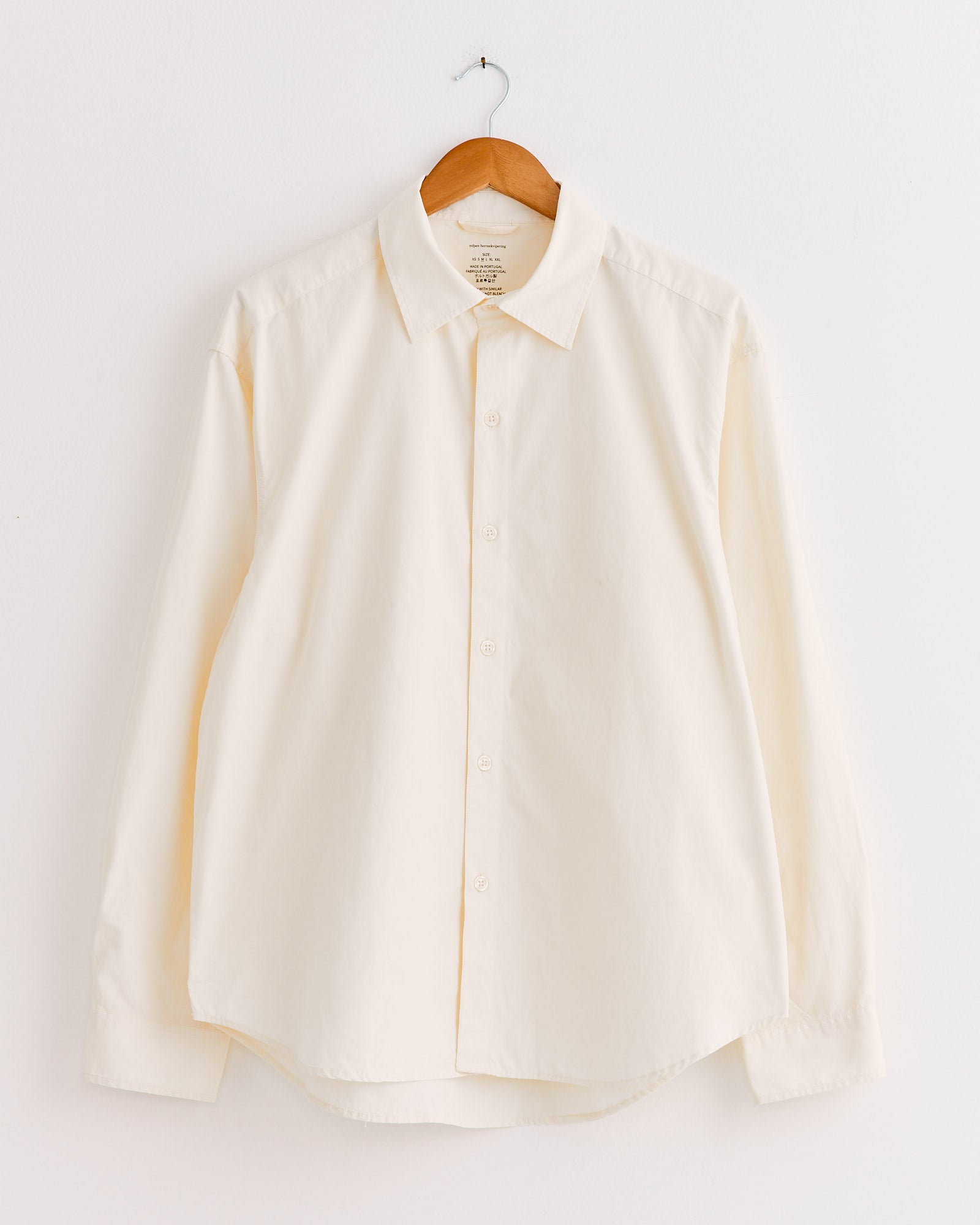 The mfpen Generous Shirt in Warm White, a relaxed fit long-sleeved button-up, hangs on a wooden hanger against a white wall.