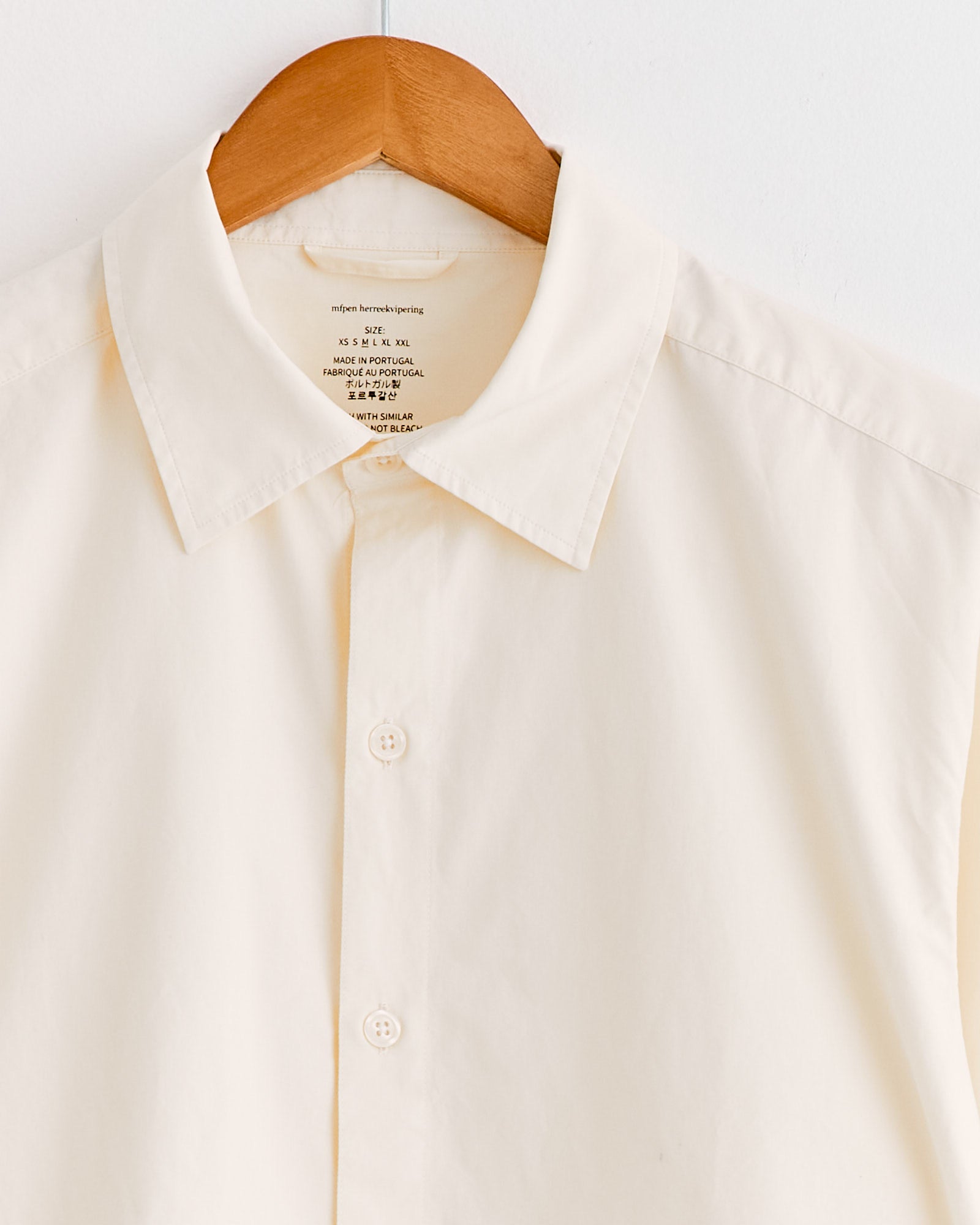 The mfpen Generous Shirt in Warm White hangs on a wooden hanger against a white background. This relaxed-fit shirt has short sleeves, a pointed collar, and visible white buttons.