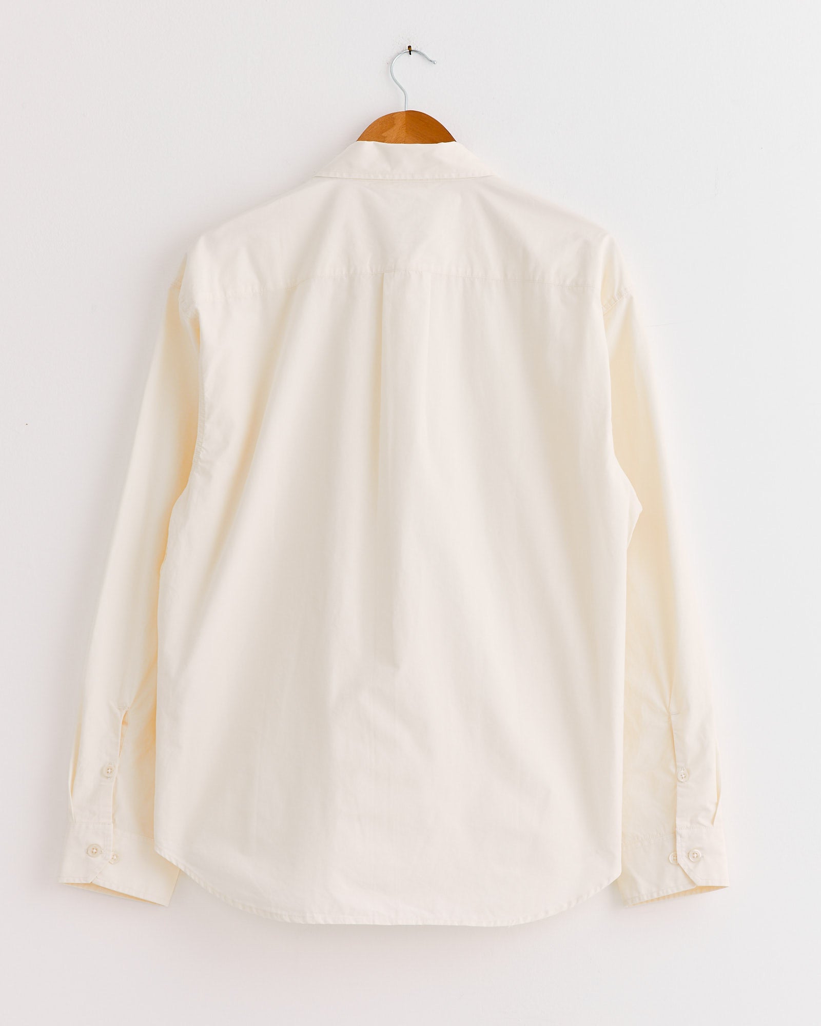 The mfpen Generous Shirt in Warm White, shown from the back on a wooden hanger against a white wall, features long sleeves and a relaxed fit that highlights its effortless style.