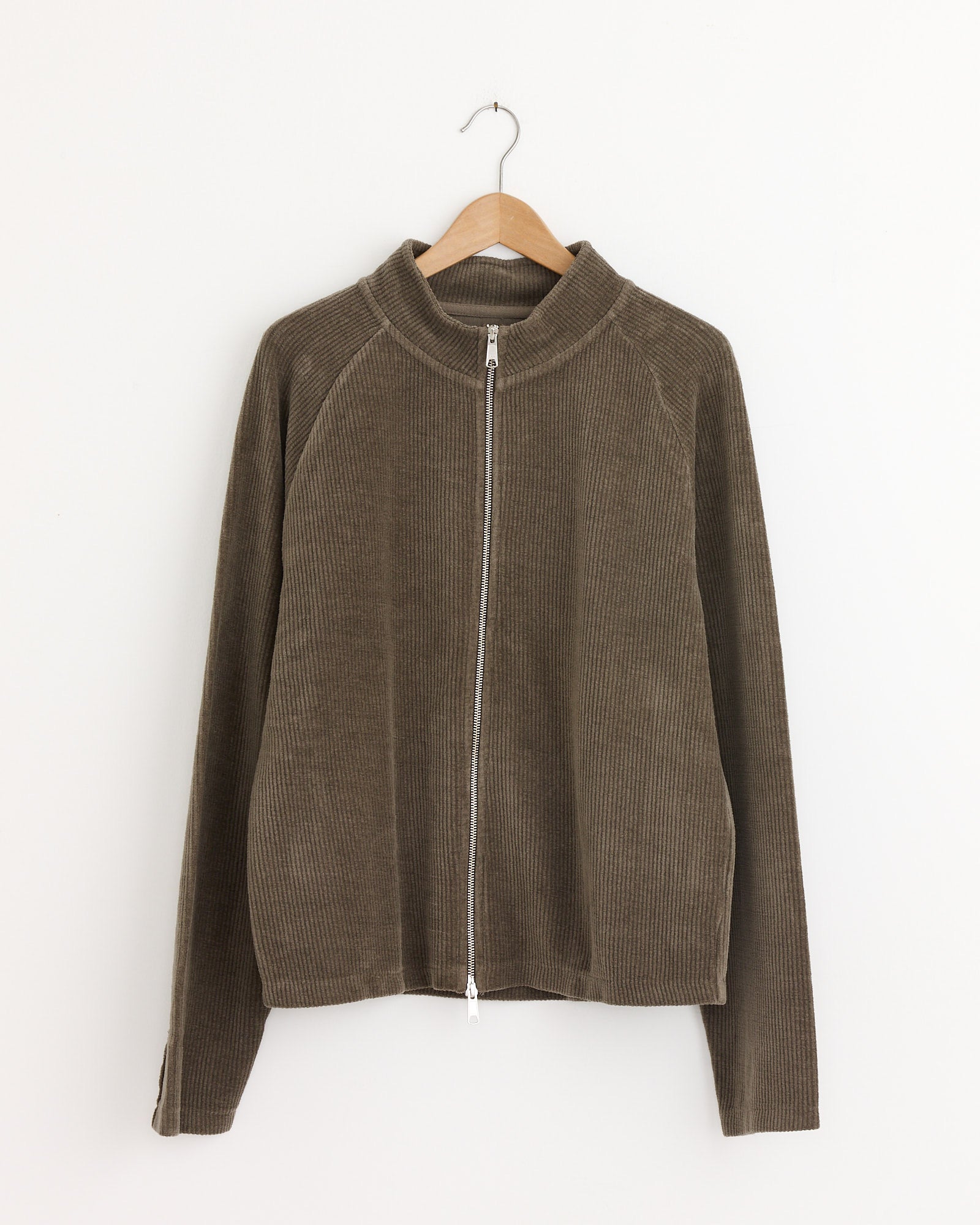 The mfpen Leisure Zip Up in Faded Taupe, a relaxed fit cotton corduroy jersey jacket with long sleeves, hangs on a wooden hanger against a plain white wall.