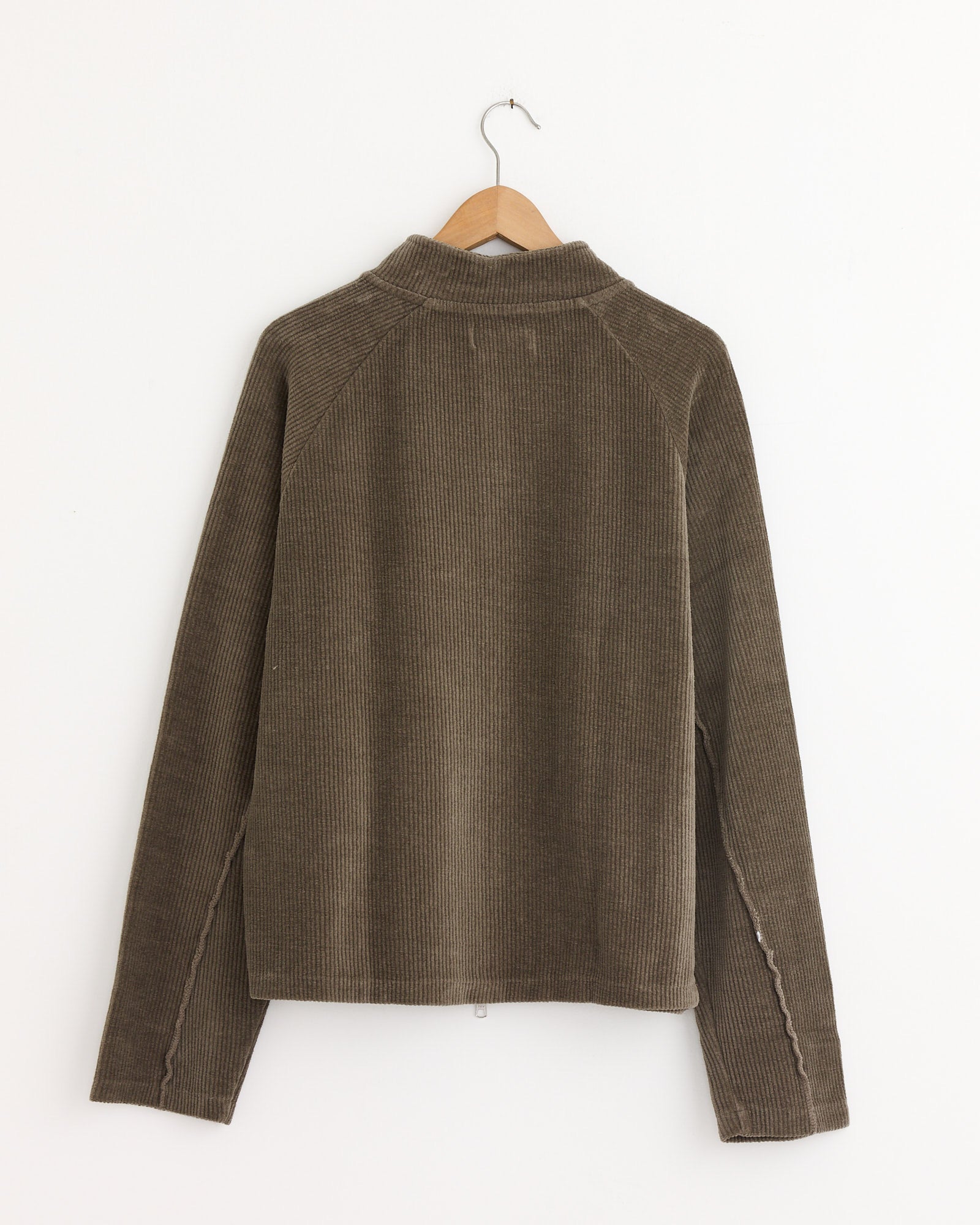 The mfpen Leisure Zip Up in Faded Taupe, featuring a relaxed fit, collar, and long sleeves, hangs on a wooden hanger against a white wall, displayed from the back.