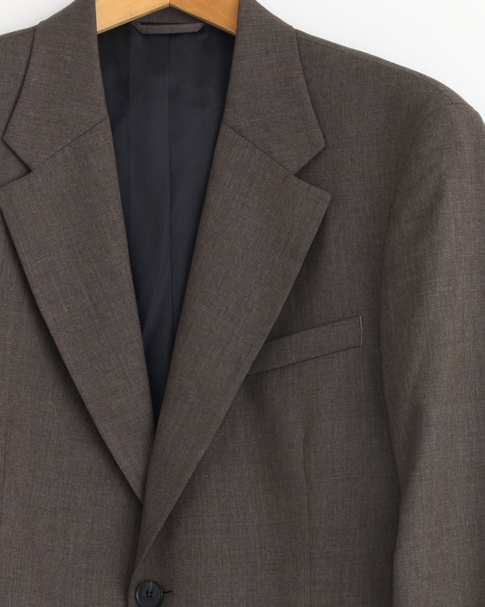A close-up of the mfpen Single Breasted Blazer in Taupe, hanging on a wooden hanger, displays the collar, lapel, button, and chest pocket of this wool twill blazer against a white background.