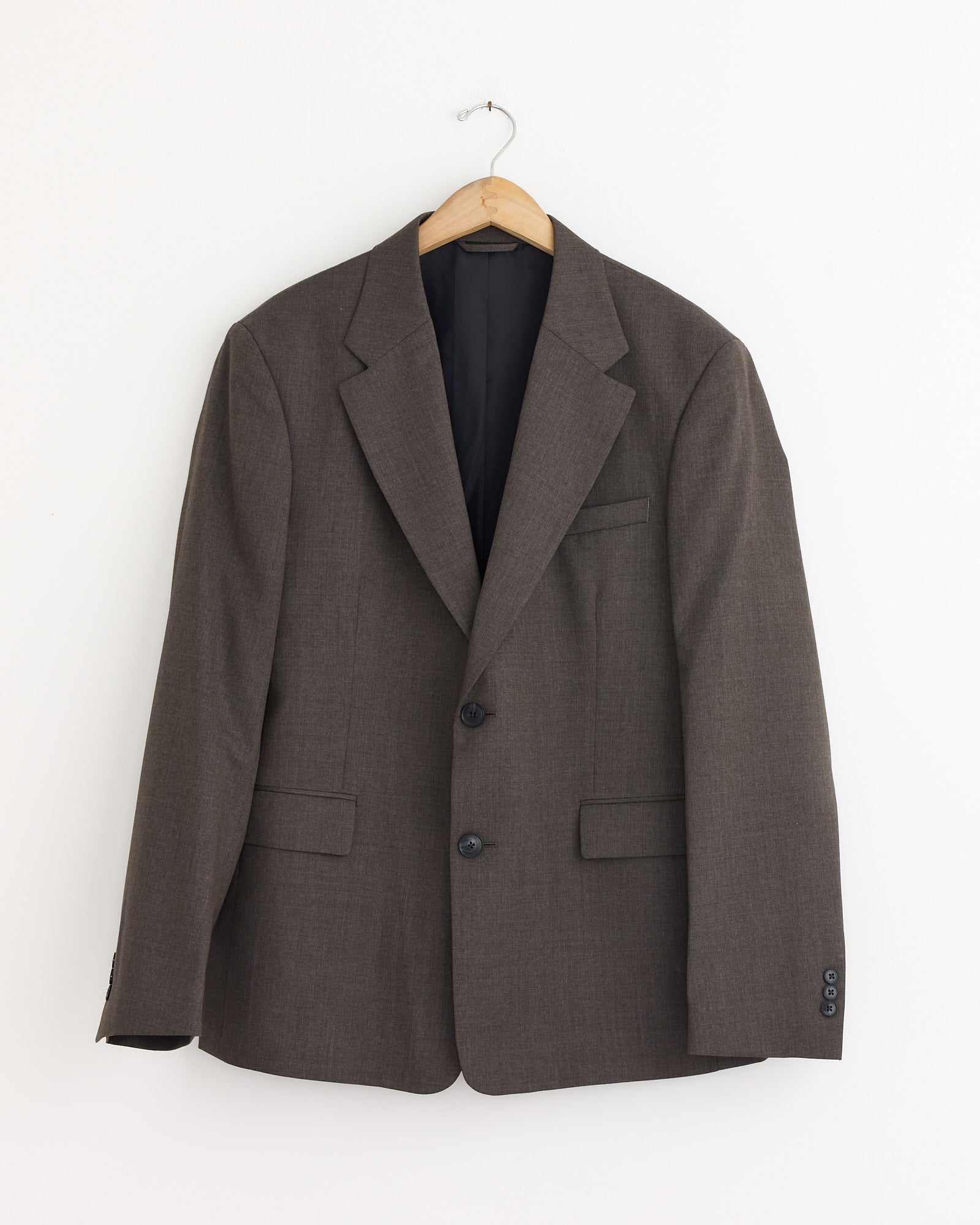 The mfpen Single Breasted Blazer in Taupe, featuring a classic two-button design and three pockets, hangs on a wooden hanger against a plain white wall.
