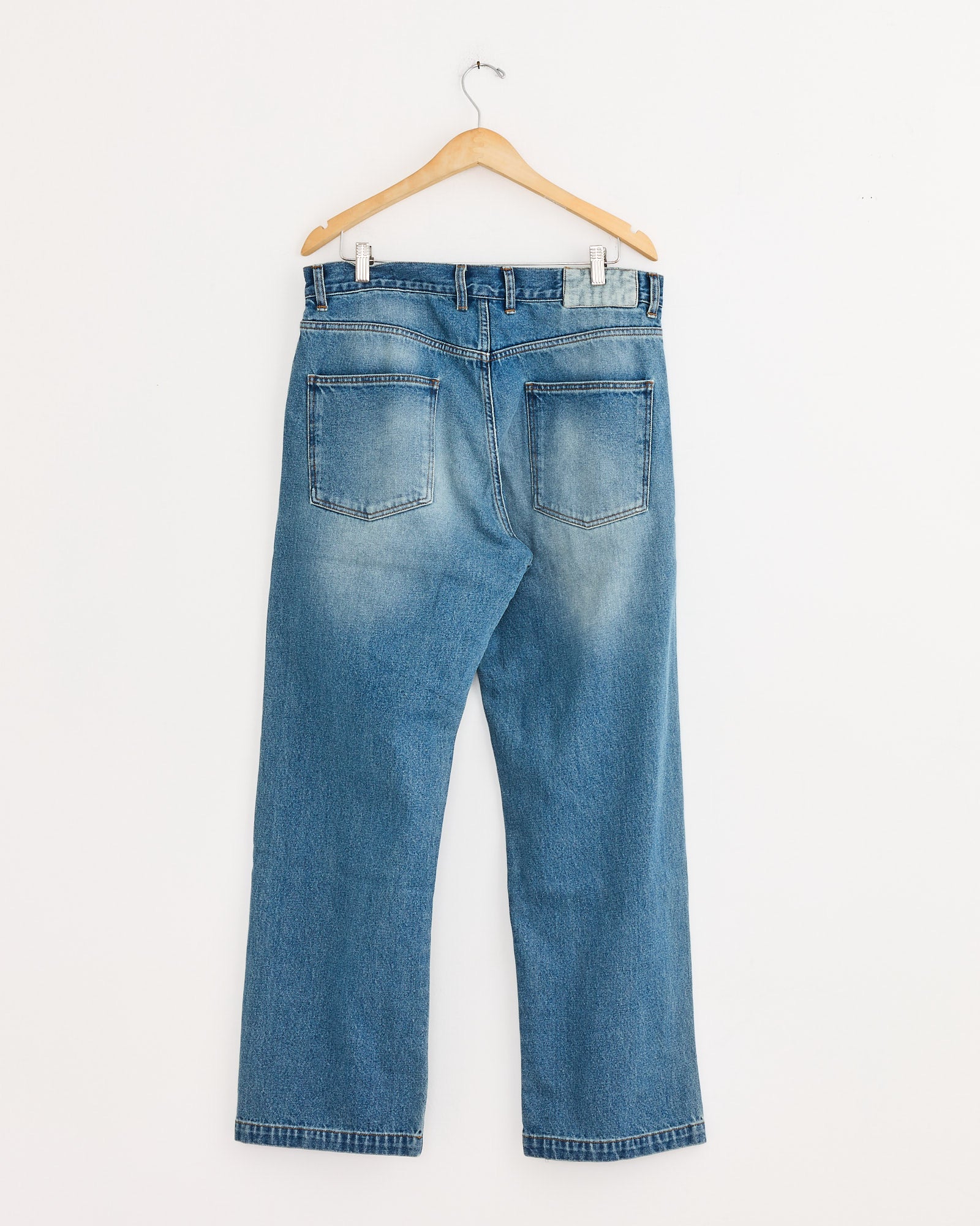 The mfpen Straight Cut Jean in Vintage Blue hangs on a wooden hanger against a plain white wall, displayed from the back with two visible rear pockets.