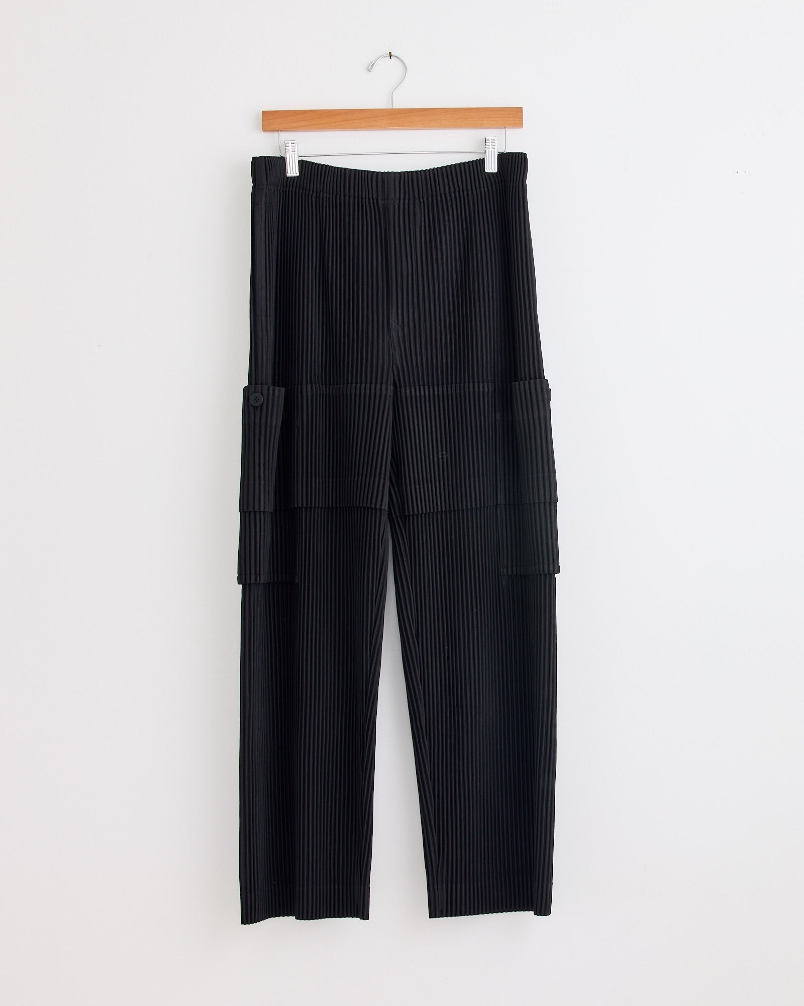 The Homme Plissé Issey Miyake Multi Pocket Pant in Coke Grey features a high waist, wide legs, and a pleated texture—perfect for any look. Shown on a wooden hanger against a white wall. Chat online for sizing help.