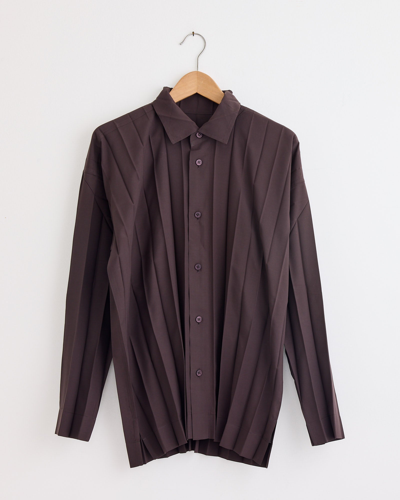 The Edge Shirt in Dark Brown by Homme Plissé Issey Miyake features long sleeves, a pointed collar, vertical pleats, and button-up front—shown on a wooden hanger against a white wall. Chat with us online for fit or sizing questions.