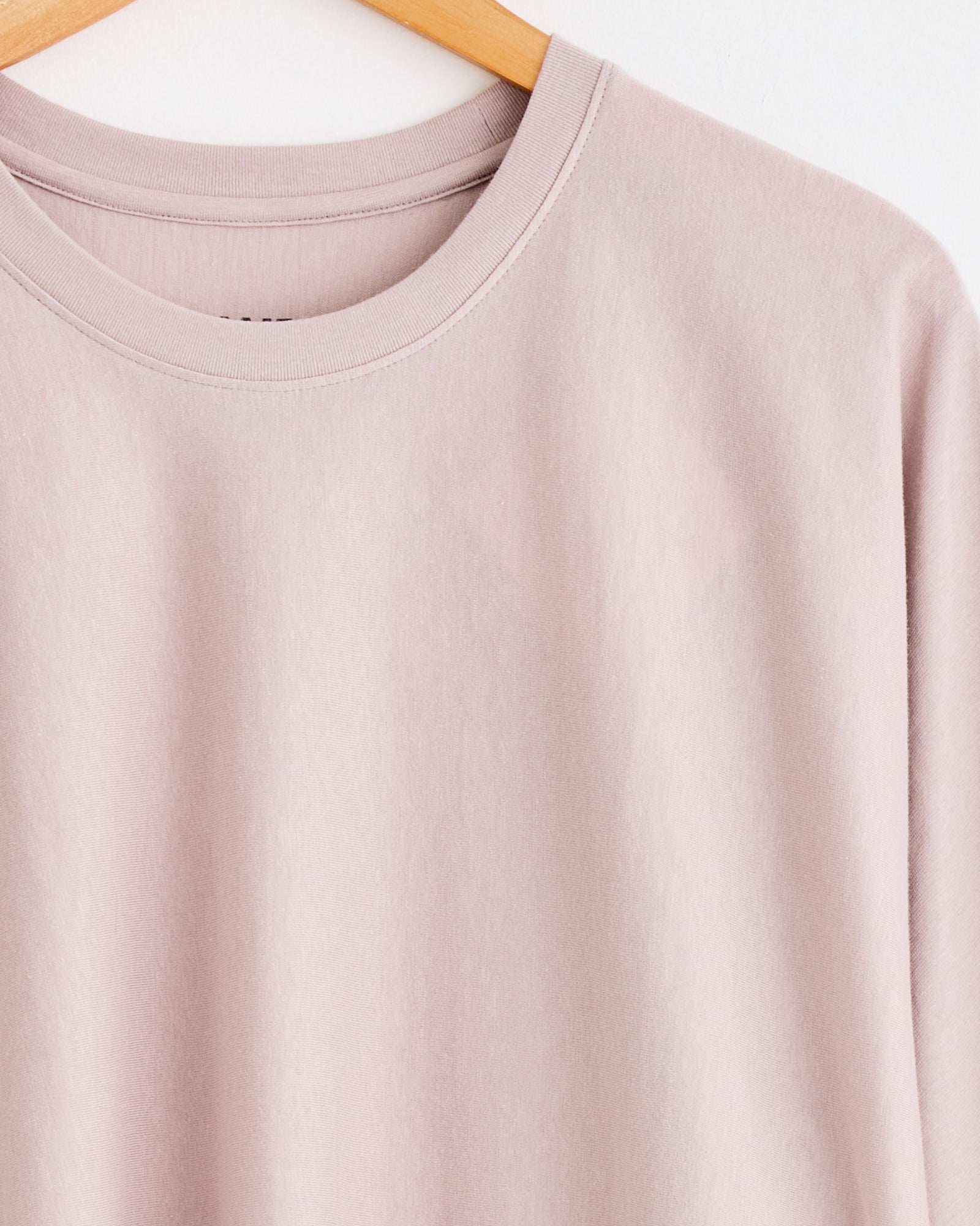 A close-up of Homme Plissé Issey Miyake's Release T1 T-Shirt in Purple Grey, focusing on its fit, hangs on a wooden hanger against a white background, with only the upper part of the shirt visible.