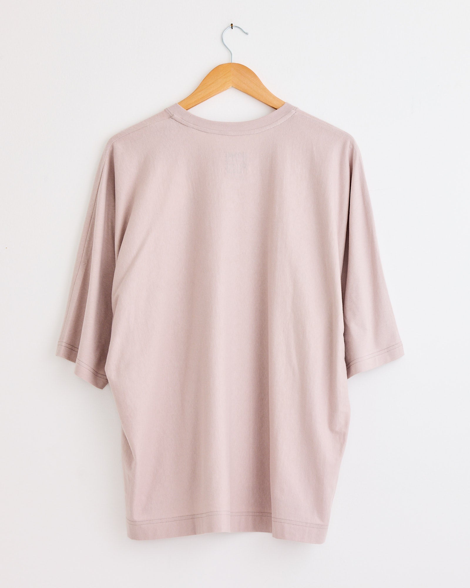 The Release T1 T-Shirt in Purple Grey by Homme Plissé Issey Miyake hangs on a wooden hanger against a white wall, shown from the back. For fit or sizing questions, please use our online chat for quick help.