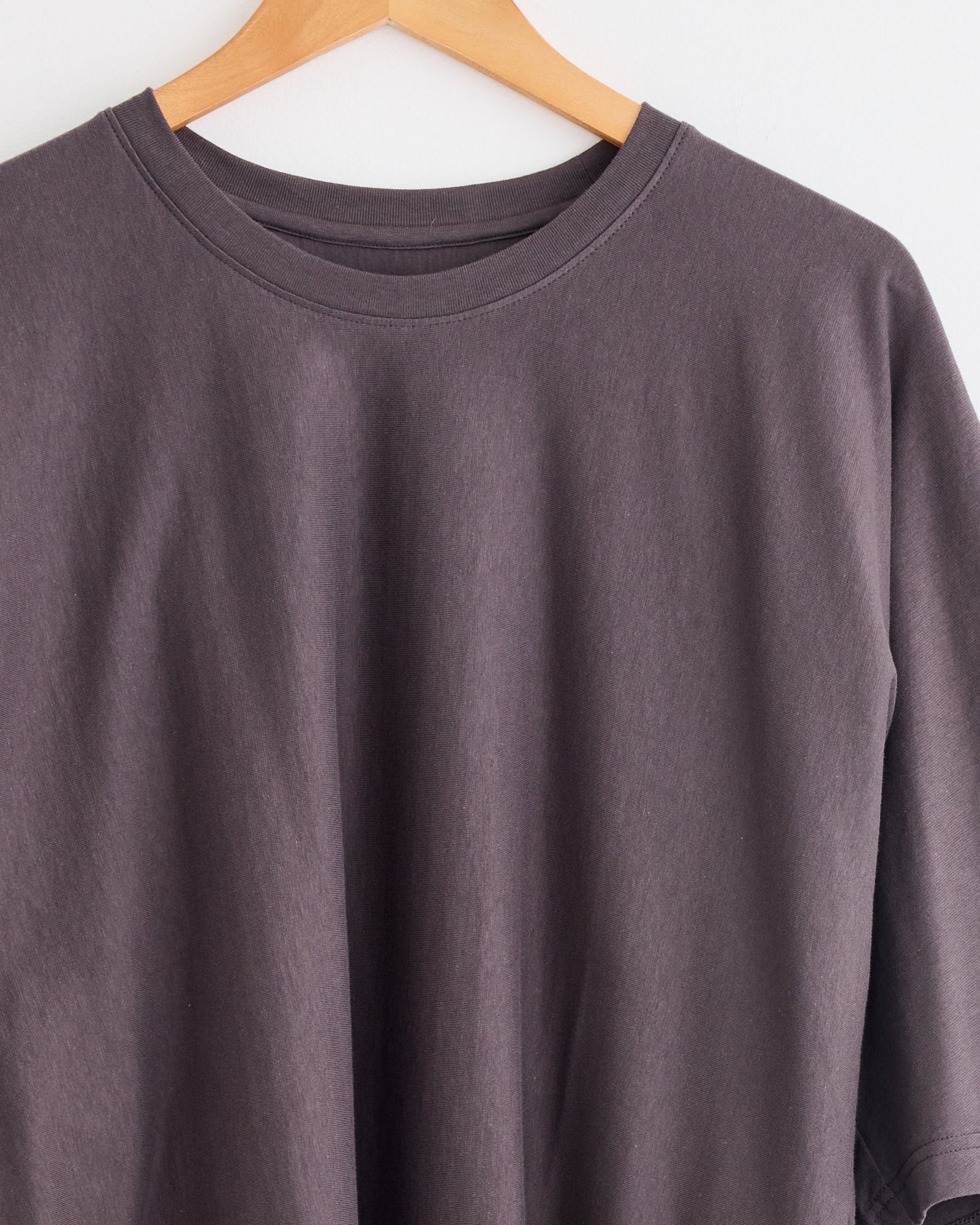 The Release T1 T-Shirt in Grey by Homme Plissé Issey Miyake hangs on a wooden hanger against a white wall—an essential for any wardrobe. For fit or sizing help, connect with our online chat to find your perfect option.