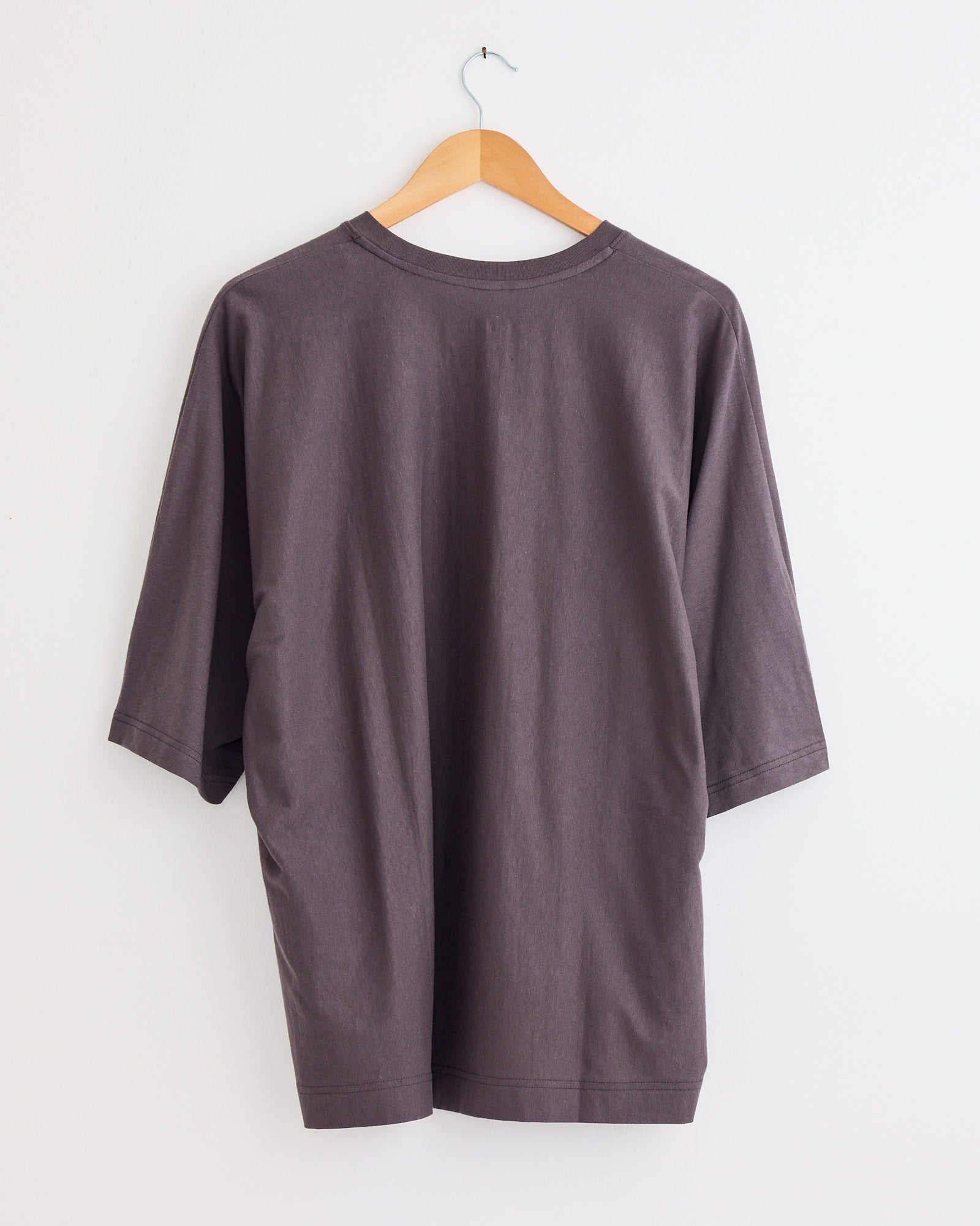 The Release T1 T-Shirt in Grey by Homme Plissé Issey Miyake hangs on a wooden hanger against a white background, displayed from the back. For fit or sizing questions, please contact us via our online chat.