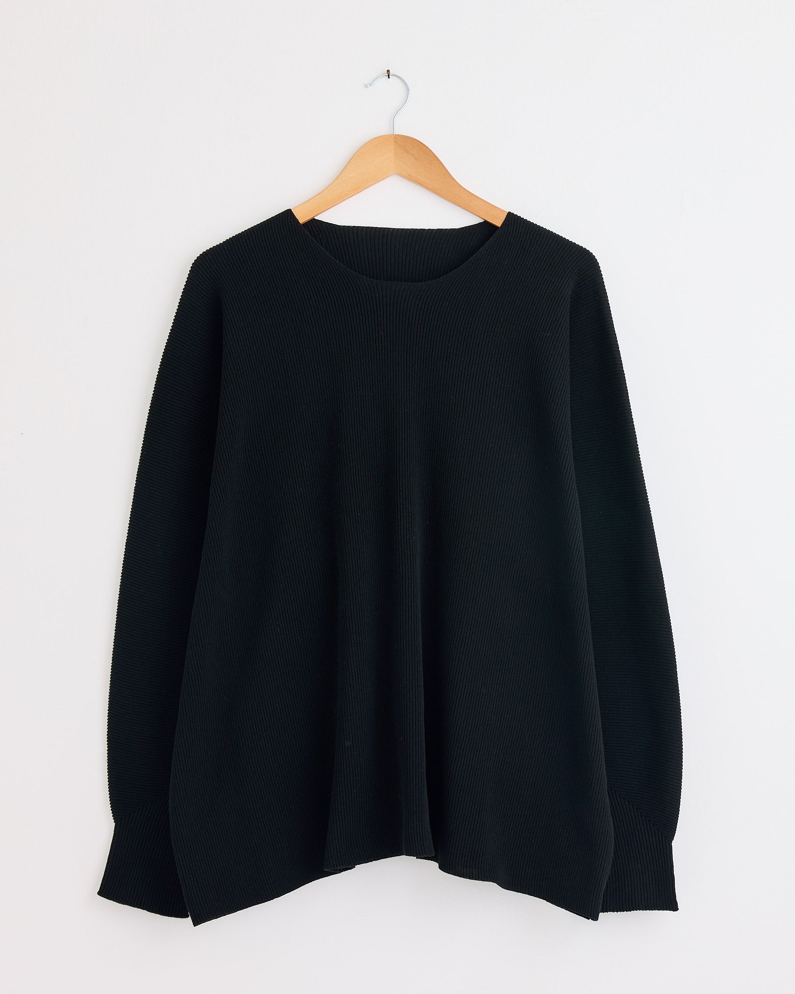 The Homme Plissé Issey Miyake Basic N280 Knit in Black, a loose-fitting long-sleeve sweater, hangs on a wooden hanger against a white wall. For sizing help, please use our online chat.