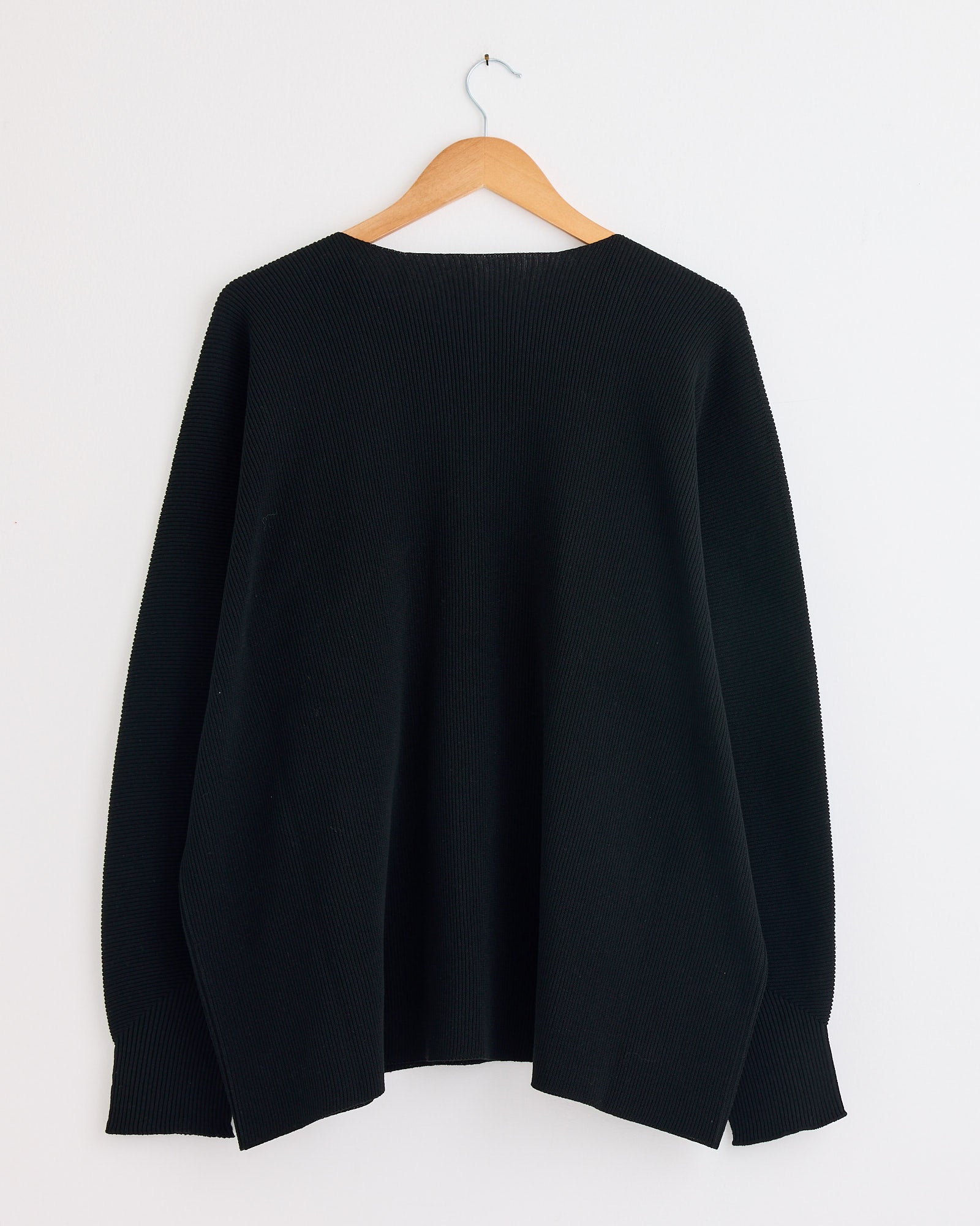 The Homme Plissé Issey Miyake Basic N280 Knit in Black, a long-sleeve sweater, hangs on a wooden hanger against a plain white wall. For sizing or fit assistance, contact us via our online chat.