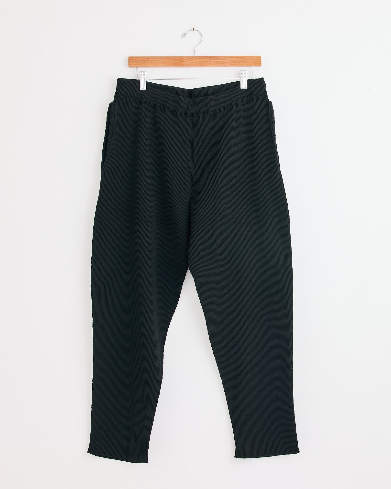 The Basic Knit Pant in Black by Homme Plissé Issey Miyake hangs on a wooden hanger against a white wall—contact our online chat for help with sizing and fit.