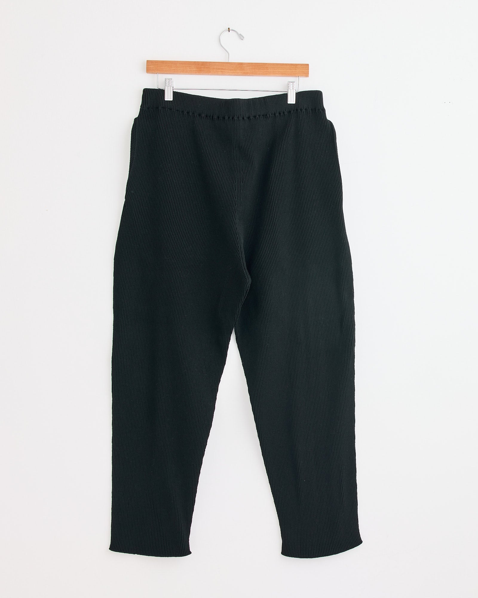 The Basic Knit Pant in Black by Homme Plissé Issey Miyake hangs on a wooden hanger against a white wall. It features a relaxed, straight-leg fit with an elastic waistband. For sizing questions, contact our online chat team.