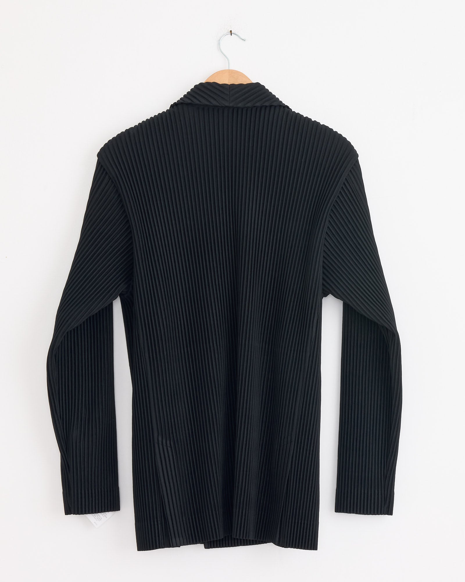 The Tailored Pleats 1 Blazer in Black by Homme Plissé Issey Miyake hangs on a wooden hanger against a white wall, shown from the back with vertical pleats and a folded collar.