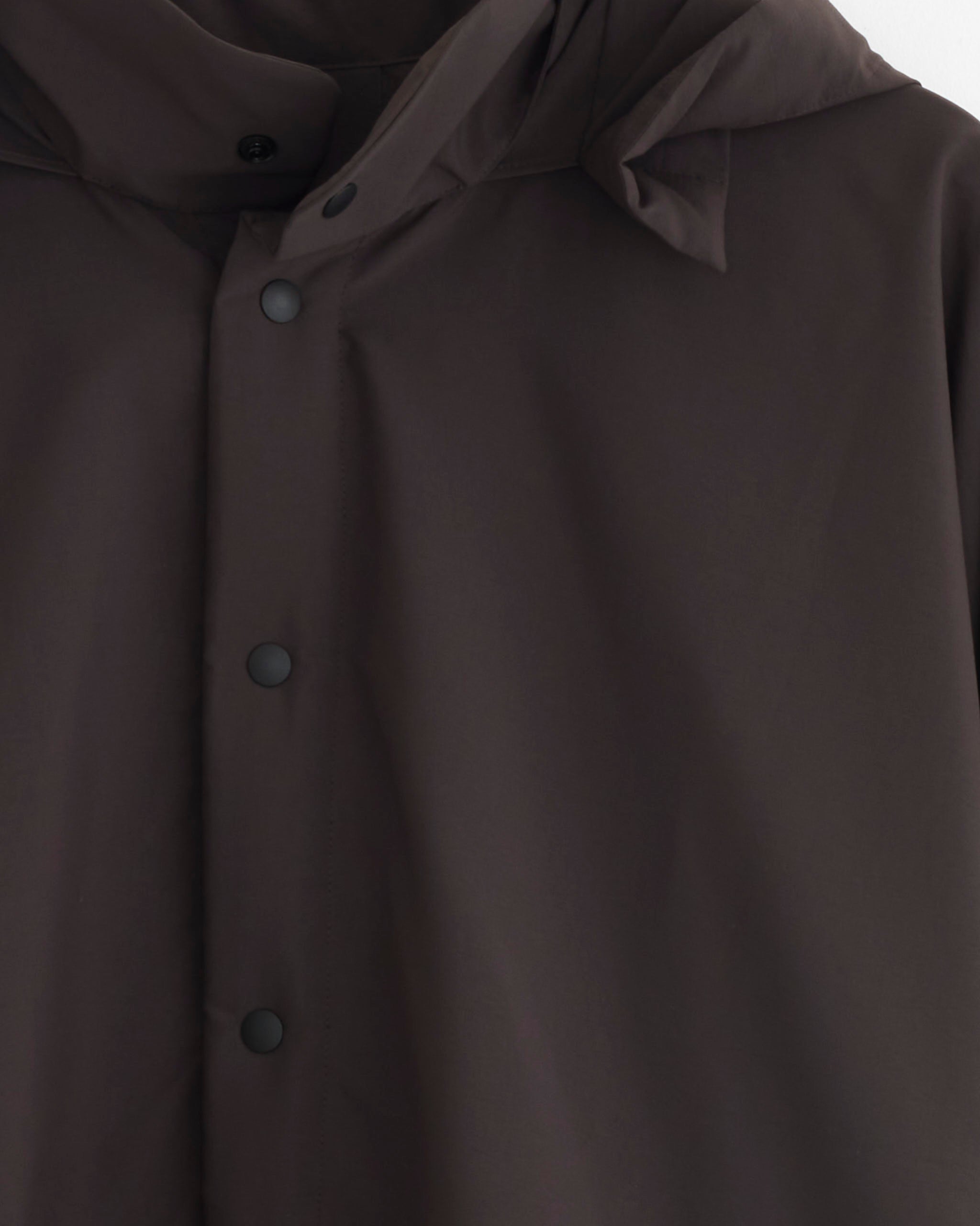 Close-up of the Padded Rim Shirt in Brown by Homme Plissé Issey Miyake, featuring a smooth, slightly reflective fabric. The focus is on the hood and chest area, with matte black snap buttons for a sleek finish.