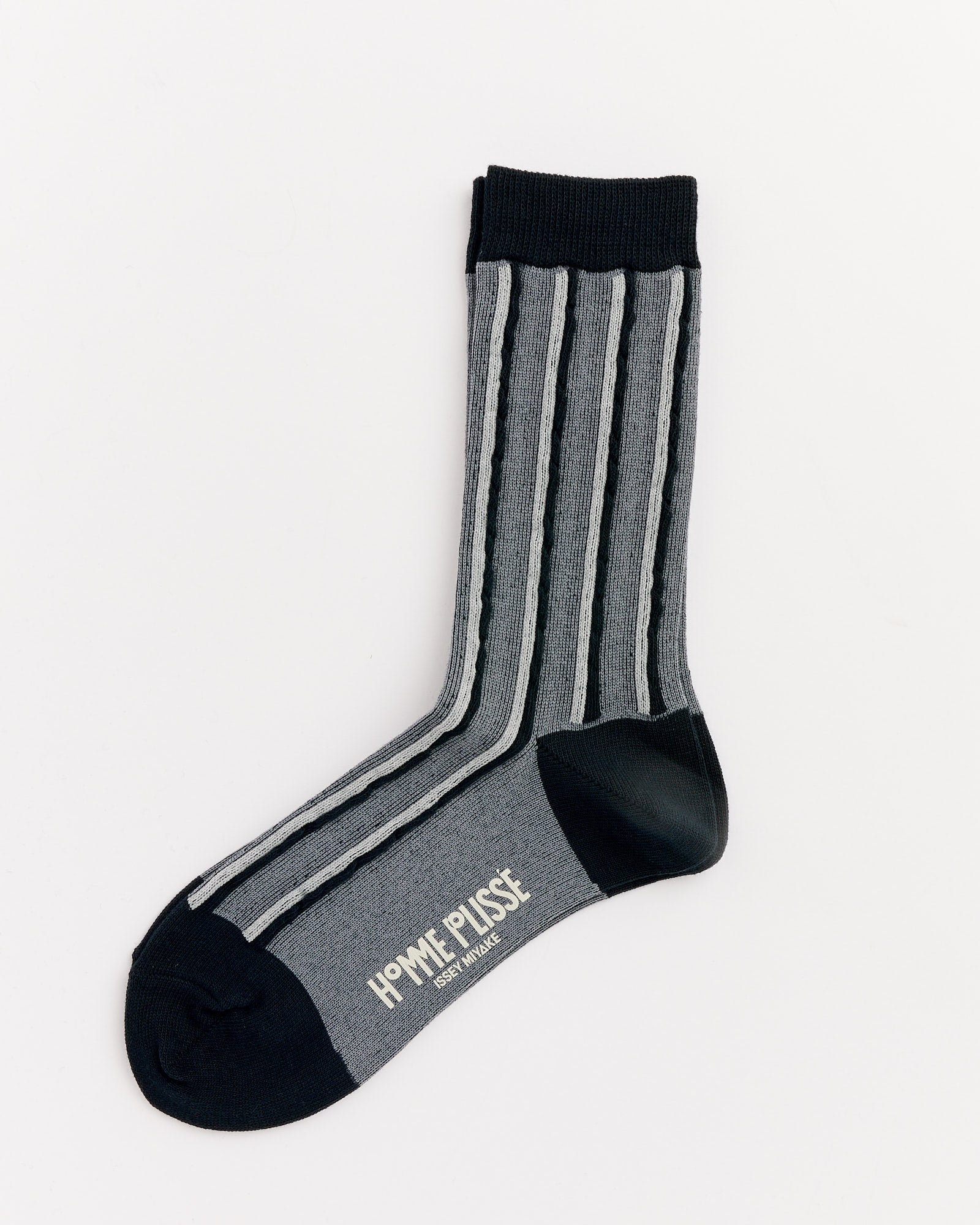 A single Masonry Sock in Black by Homme Plissé Issey Miyake, featuring gray with black and white vertical stripes, black toe, heel, and cuff. "HOMME PLISSÉ ISSEY MIYAKE" is printed in white on the sole. Made in Japan and laid flat on a white background.