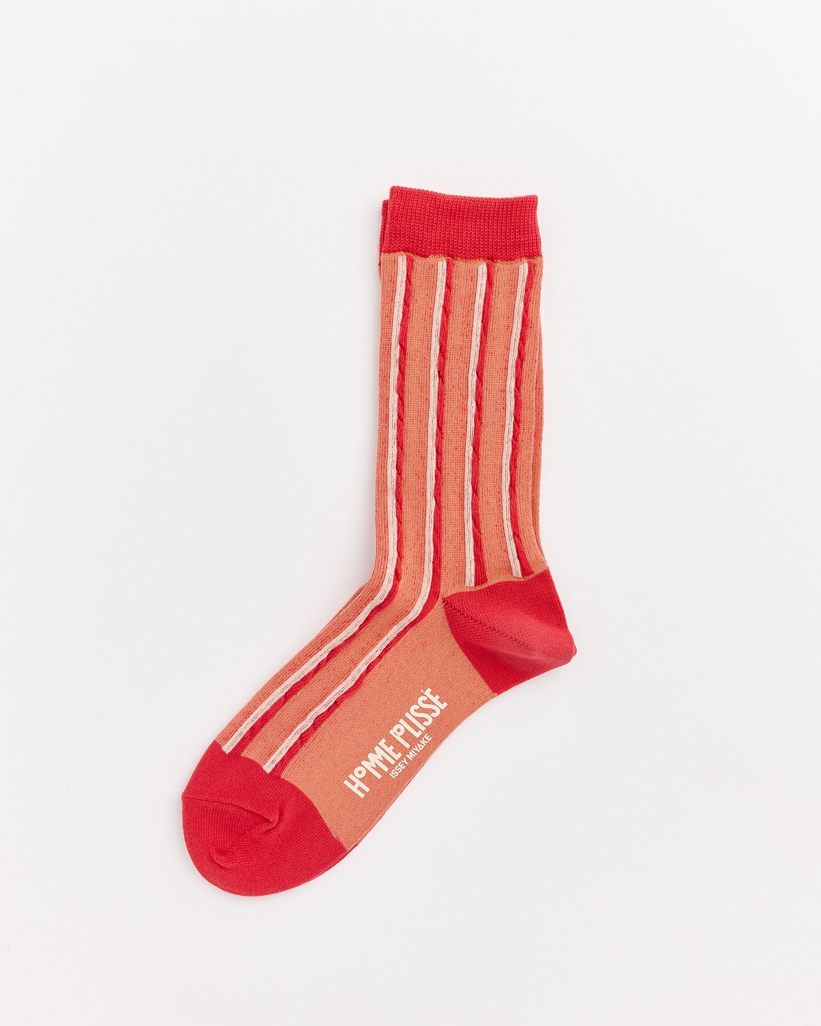 The Masonry Sock in Red by Homme Plissé Issey Miyake is crafted from Japanese cotton with beige vertical stripes, a solid red heel, toe, and cuff, and features white "HOMME PLISSÉ ISSEY MIYAKE" text on the sole.