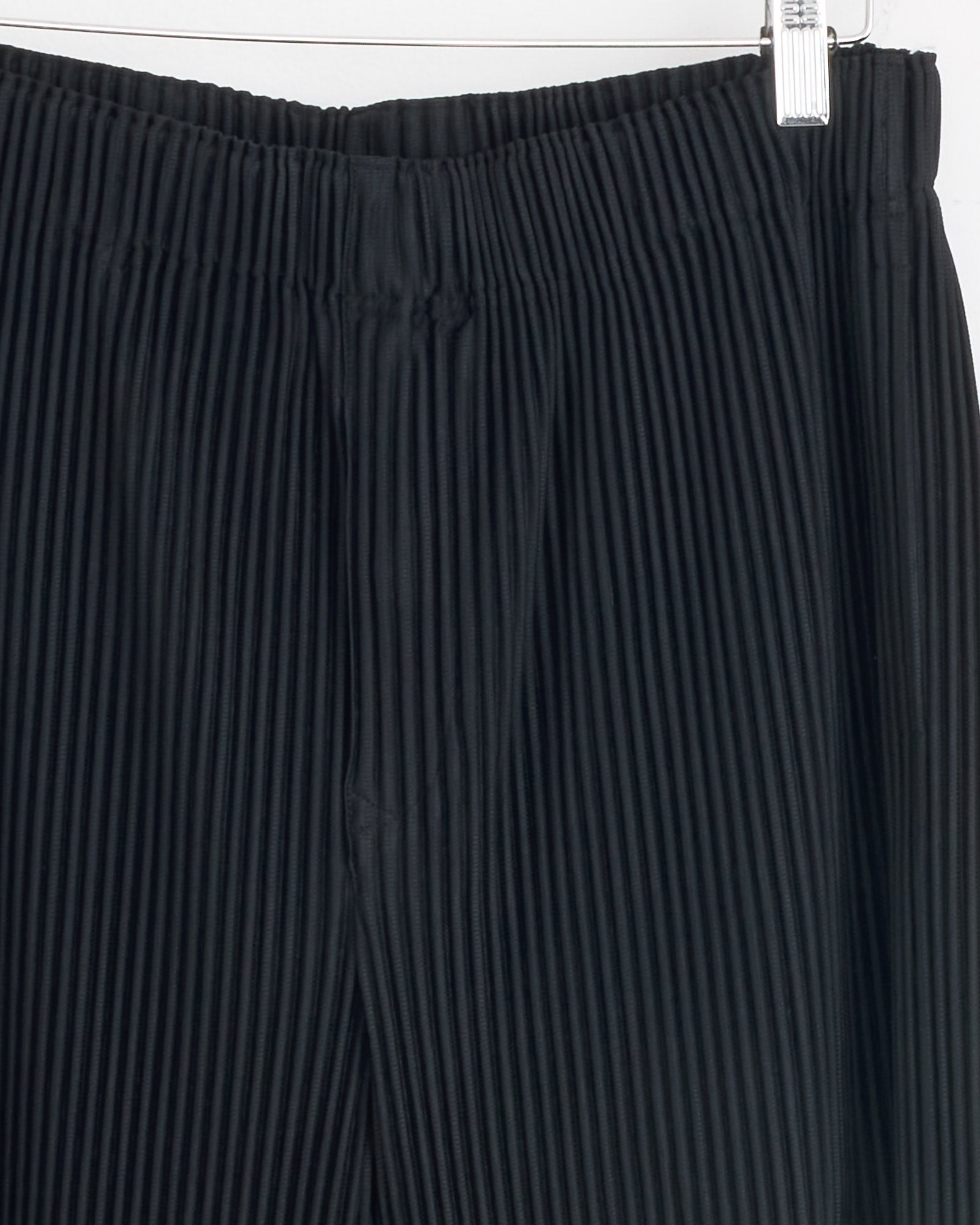Close-up of the MC Oct Pant in Black by Homme Plissé Issey Miyake, hanging on a hanger and showcasing its waistband and vertical pleats in black polyester.