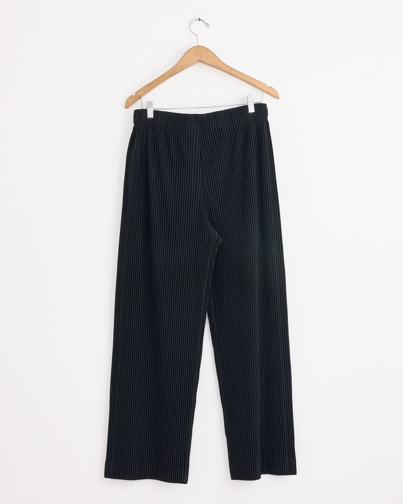 A pair of Homme Plissé Issey Miyake MC Oct Pants in black, featuring thin white vertical pinstripes, hangs on a wooden hanger against a white background.