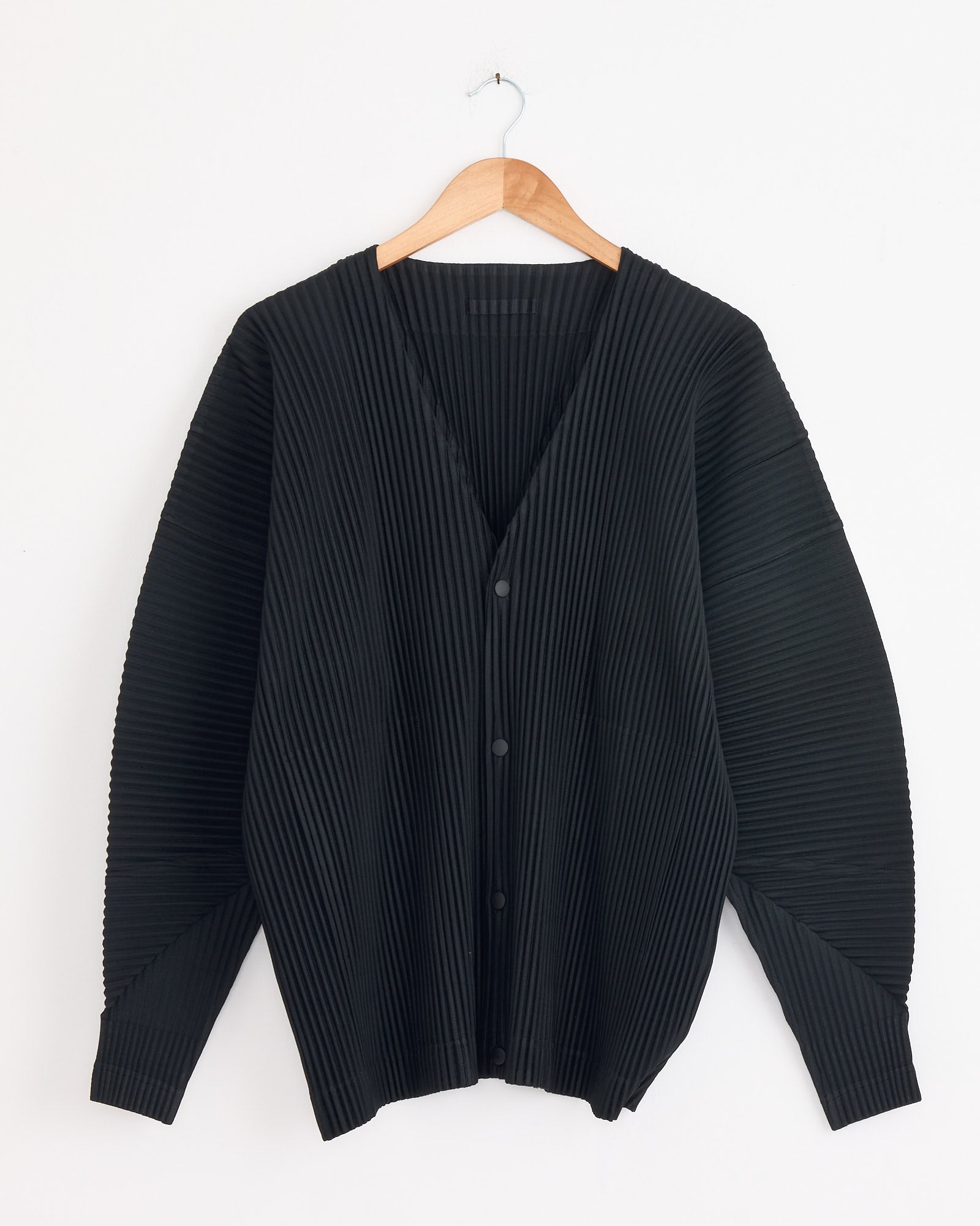 The MC Oct Cardigan in Black by Homme Plissé Issey Miyake—a black, long-sleeved, pleated V-neck cardigan with button-up front—is displayed on a wooden hanger against a white wall. For sizing questions, contact us via online chat.