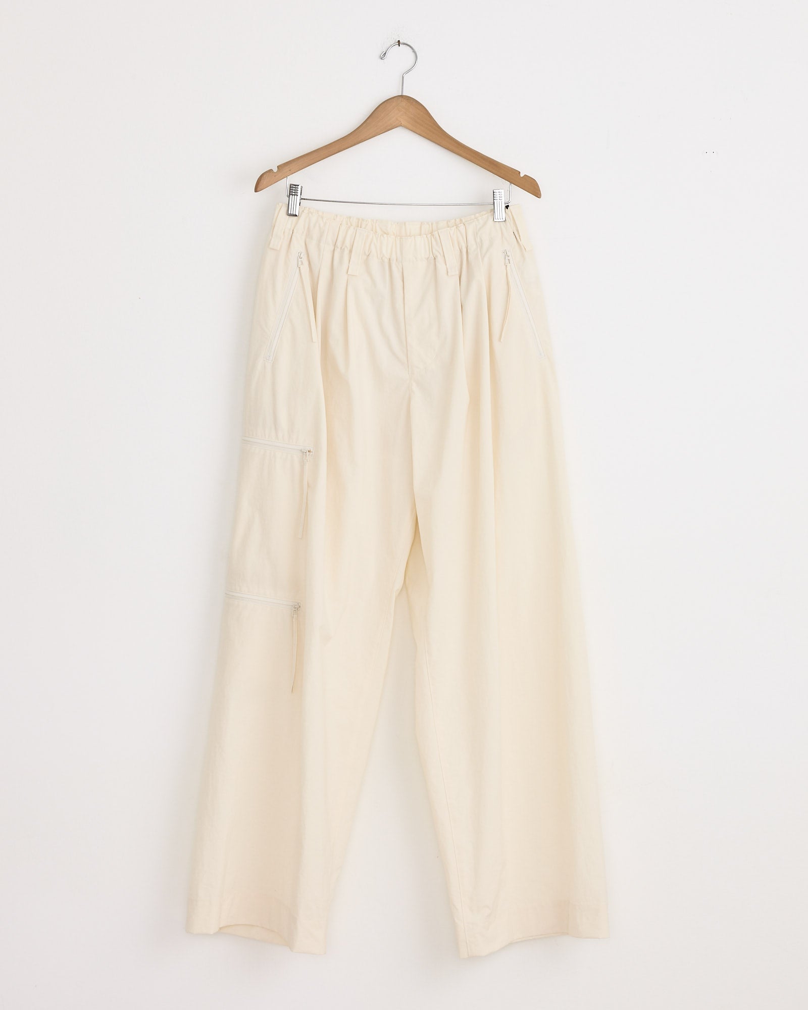 The Quarry Pant in White by Homme Plissé Issey Miyake features a loose fit, elastic waistband, side pockets, and a zipper pocket on one leg. Shown hanging on a wooden hanger against a plain white wall.