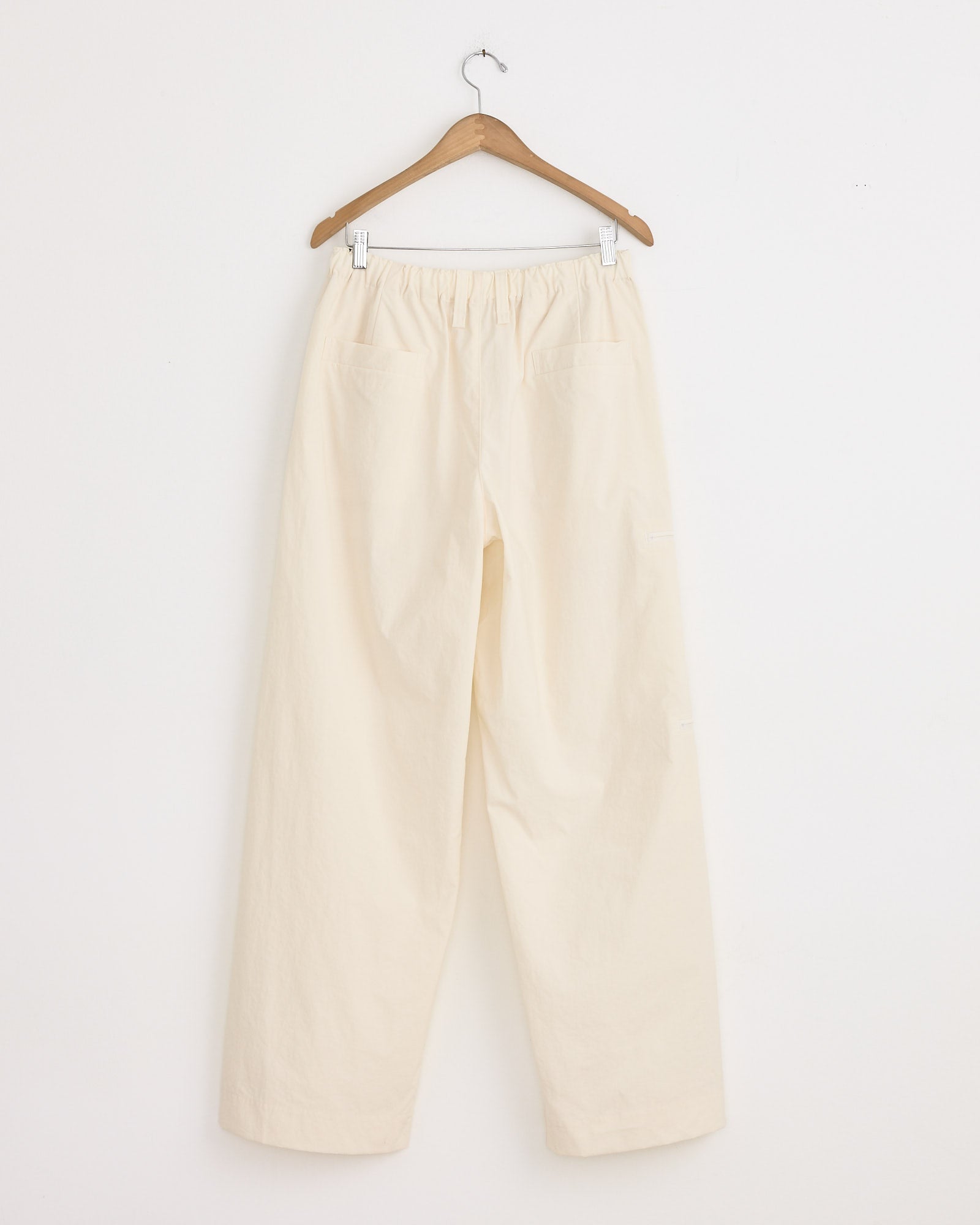 The Quarry Pant in White by Homme Plissé Issey Miyake, featuring an elastic waistband and side pockets, is shown hanging on a wooden hanger against a plain white wall.