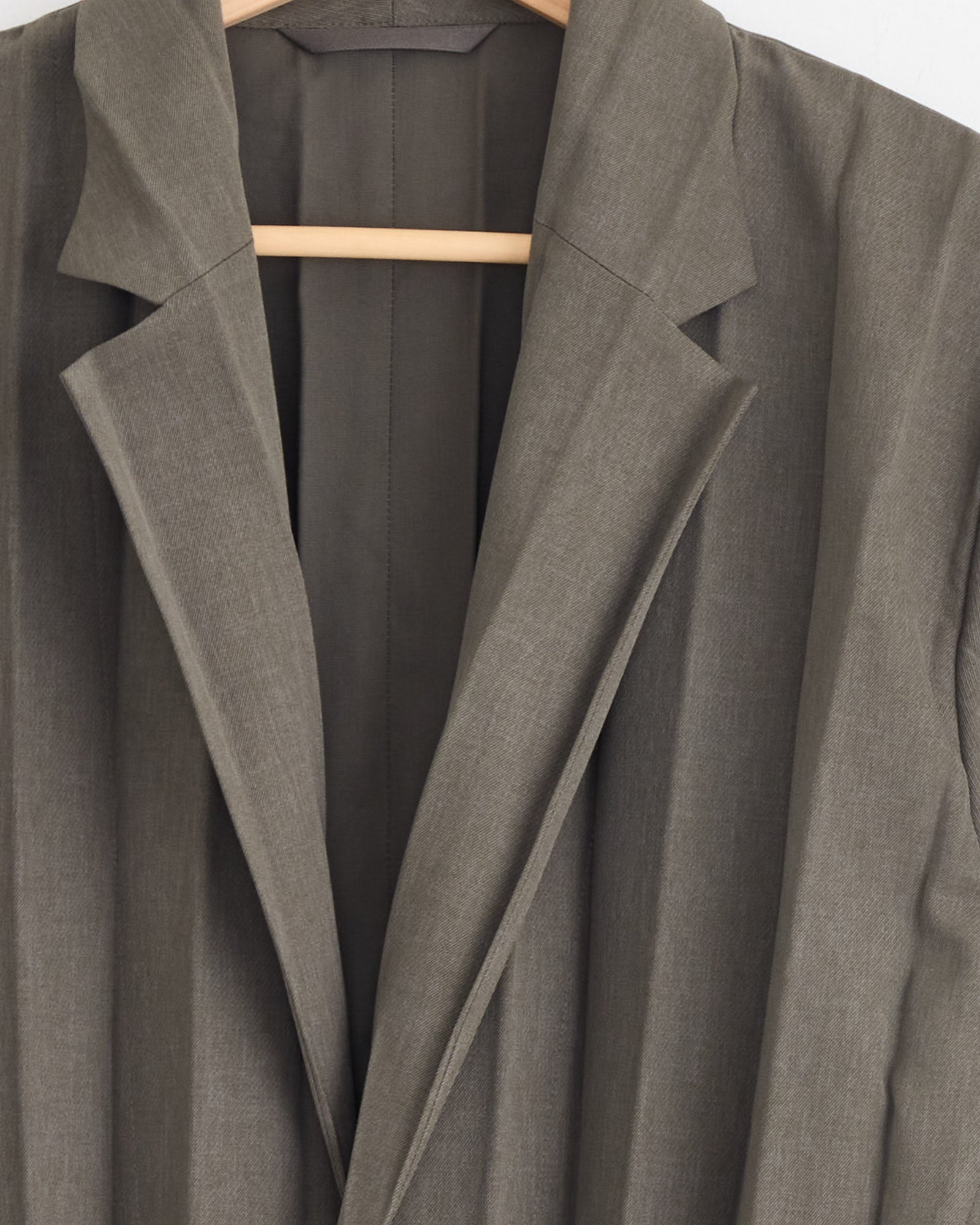 A close-up of the Edge Ensemble Blazer in Khaki by Homme Plissé Issey Miyake, showing accordion pleats on the lapels and shoulders, displayed on a wooden hanger against a plain white background.