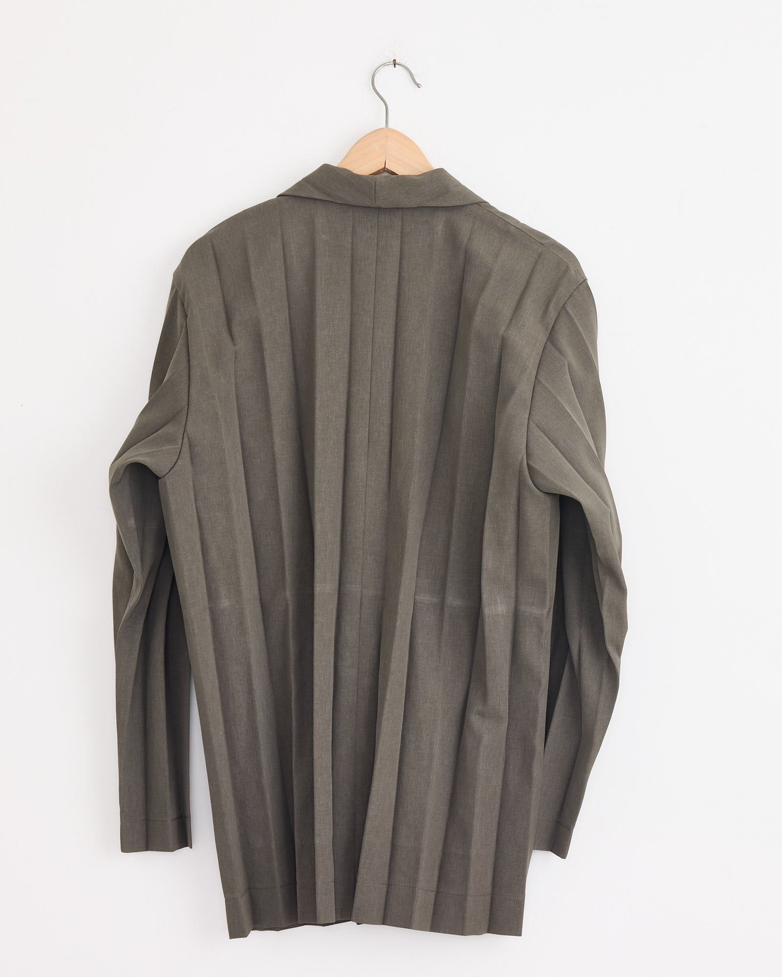 A khaki Edge Ensemble Blazer by Homme Plissé Issey Miyake hangs on a wooden hanger against a white wall, shown from the back with visible vertical seam lines and subtle accordion pleats.