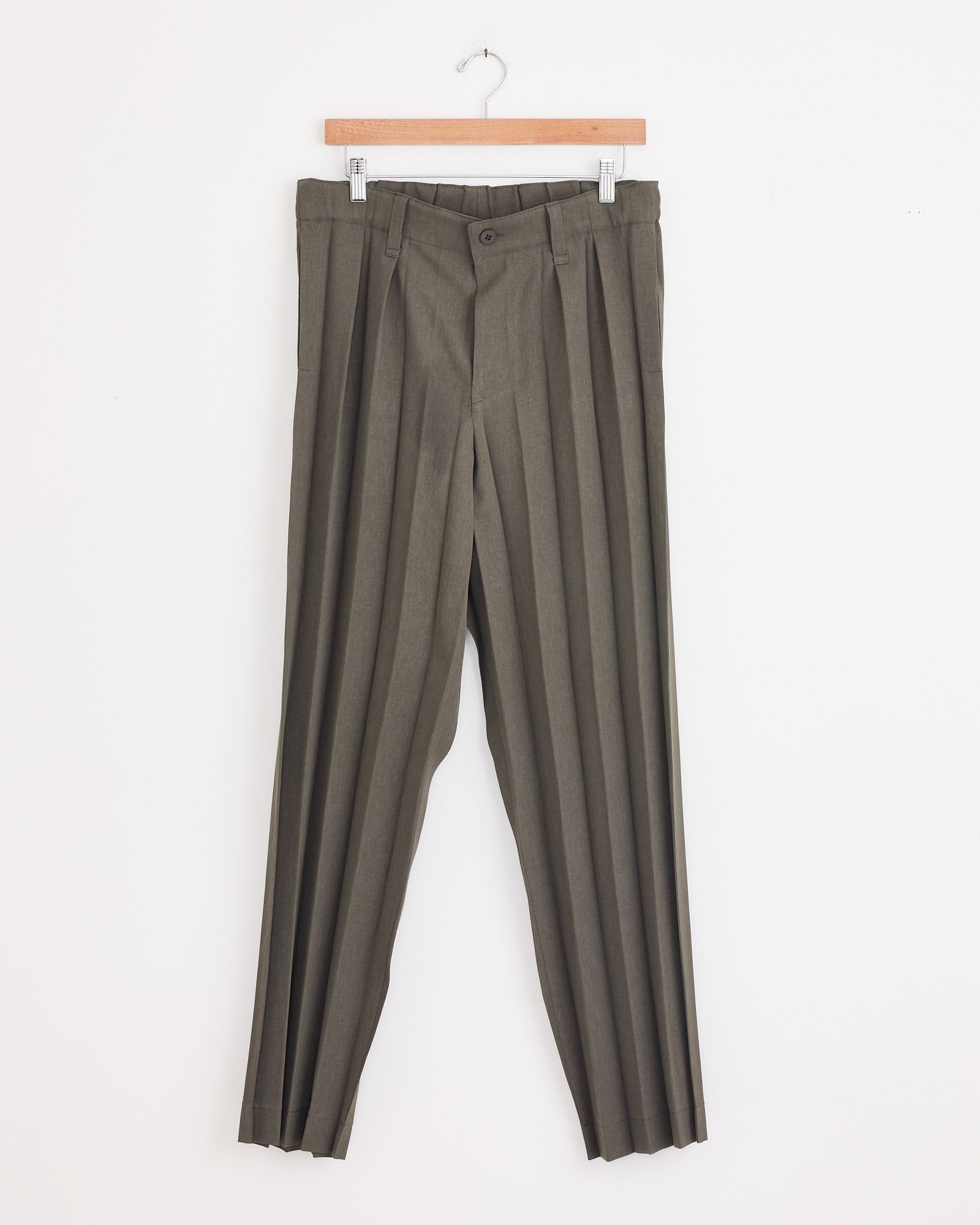 Edge Ensemble Pant in Khaki by Homme Plissé Issey Miyake with button and zipper closure, belt loops, and pronounced accordion pleats, displayed on a wooden hanger against a plain white wall.
