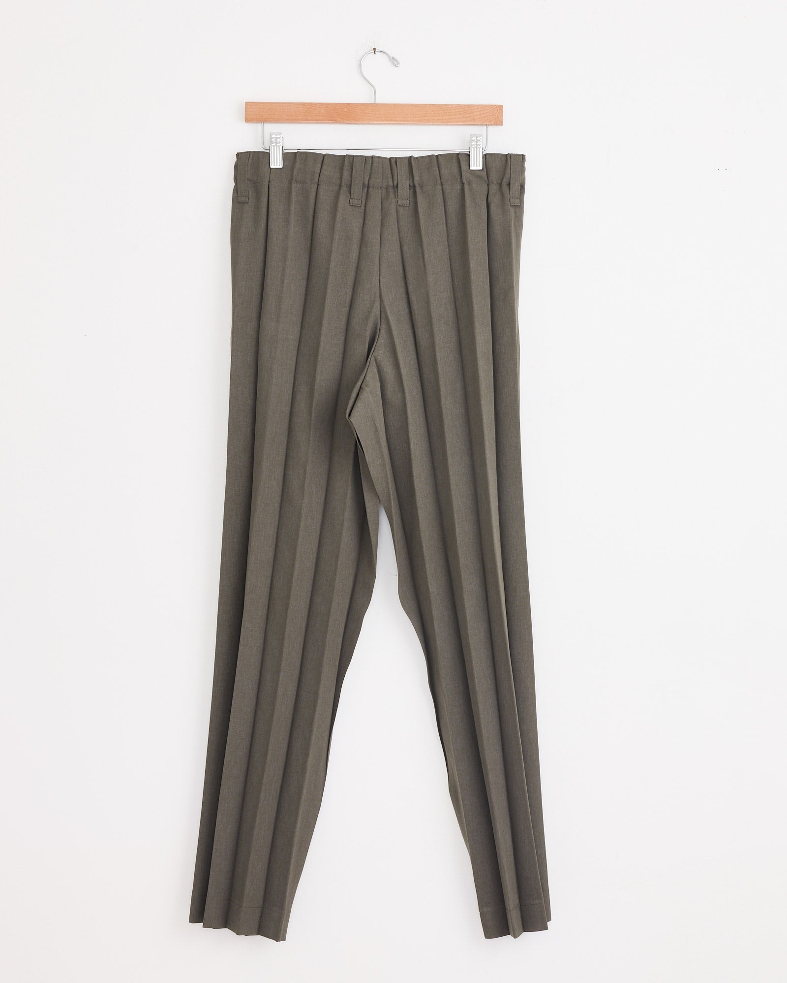 A pair of Homme Plissé Issey Miyake Edge Ensemble Pant in Khaki, featuring accordion pleats, hangs on a wooden hanger against a plain white wall.